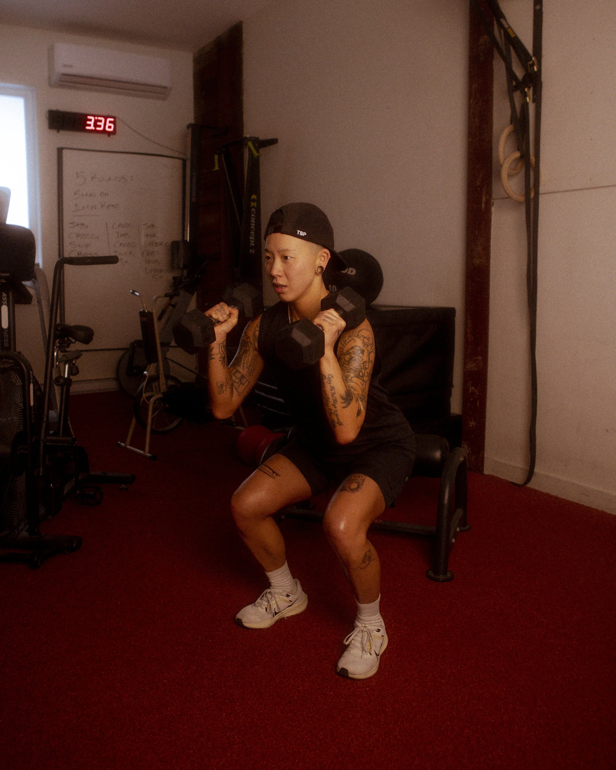 A person with tattoos and a black cap doing seated squats with dumbbells in a gym, with a whiteboard and exercise equipment in the background.