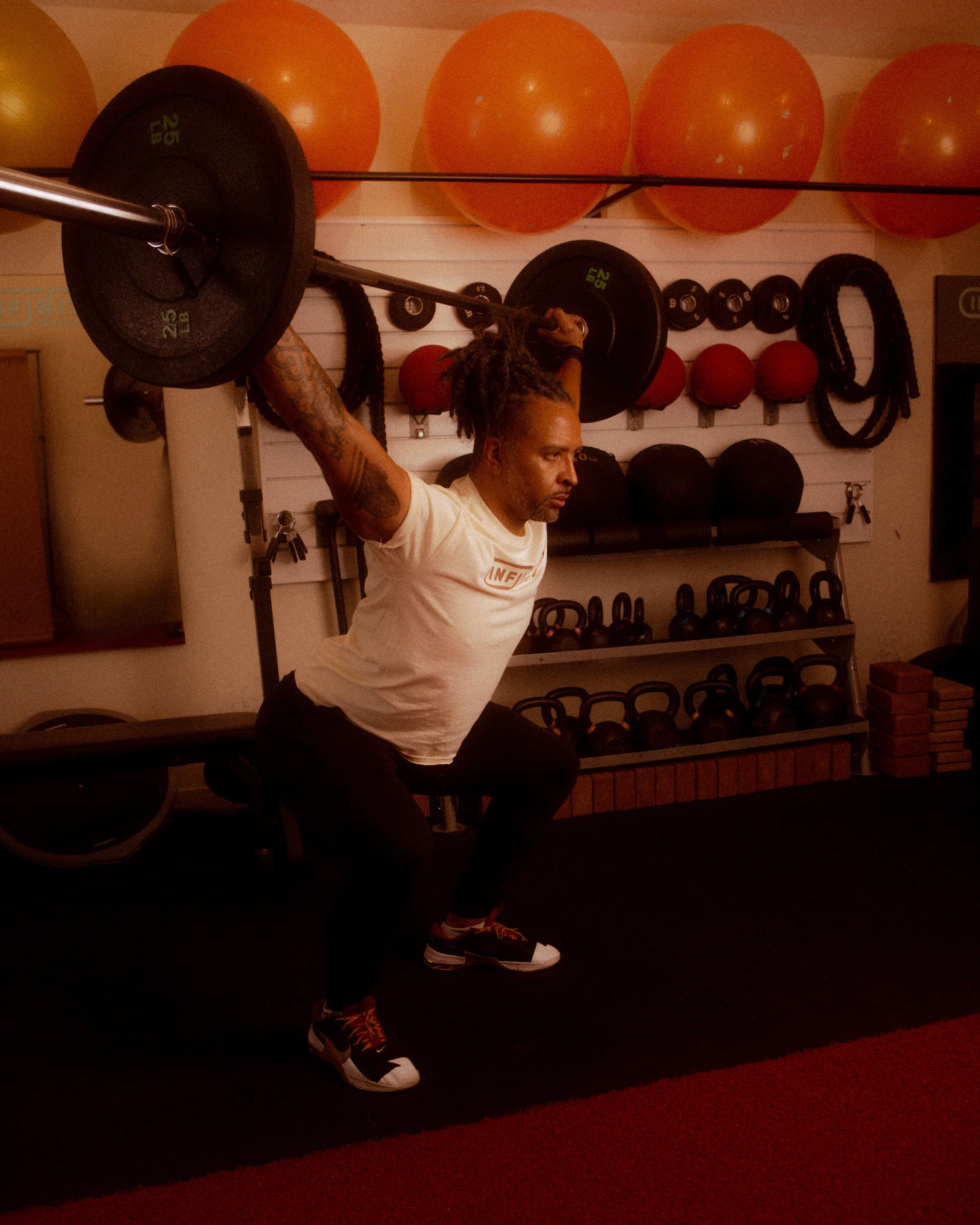 A man with dreadlocks lifting a barbell overhead during a workout in a gym with exercise balls and kettlebells in the background.