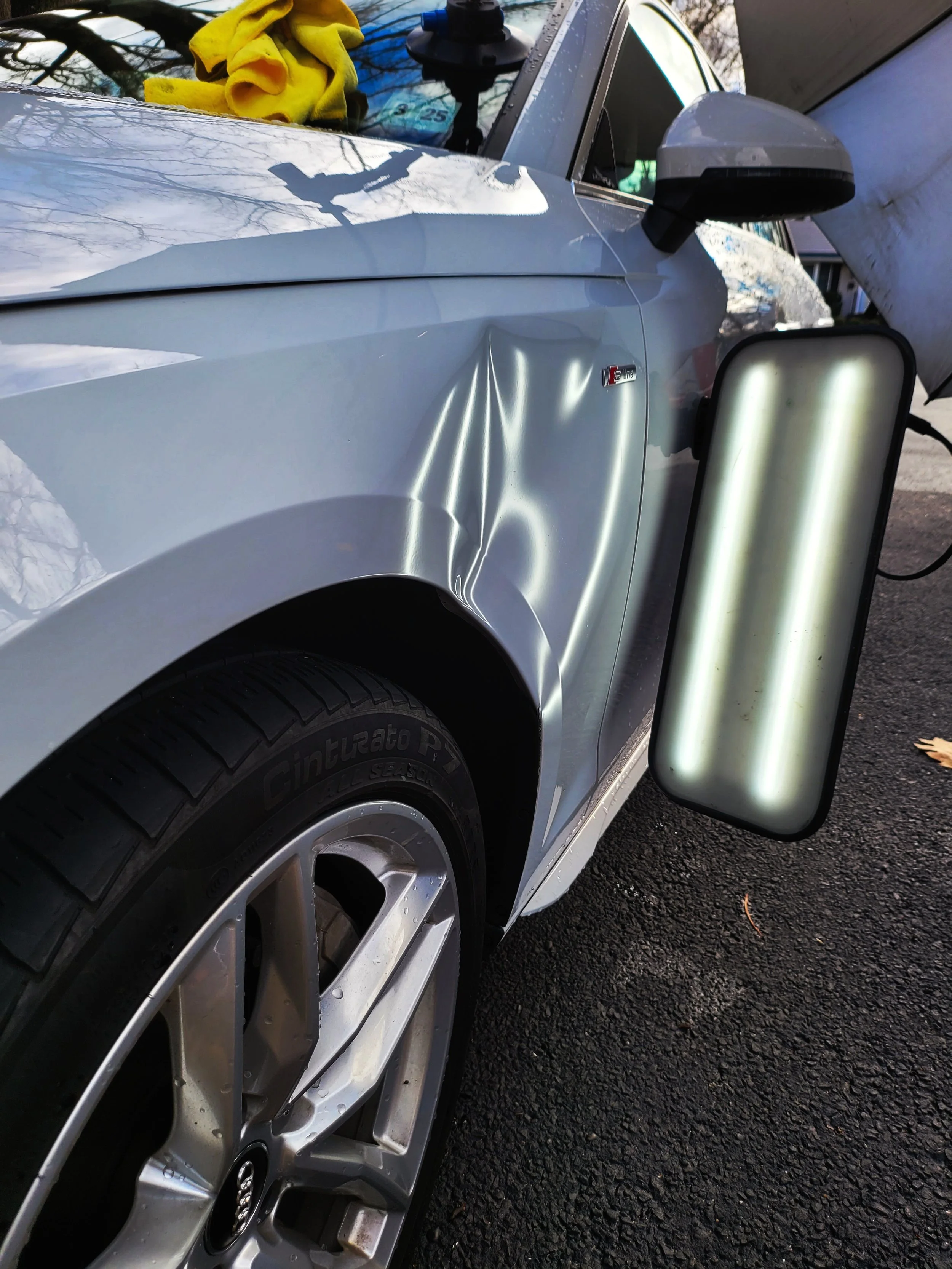 Close-up of the front left side of a silver Audi car with a large dent on the front fender. The car is parked on a paved surface, and a temporary side mirror is attached to the driver side. A yellow cloth is draped over the windshield wiper, and a large portable light is positioned beside the car, illuminating the scene.