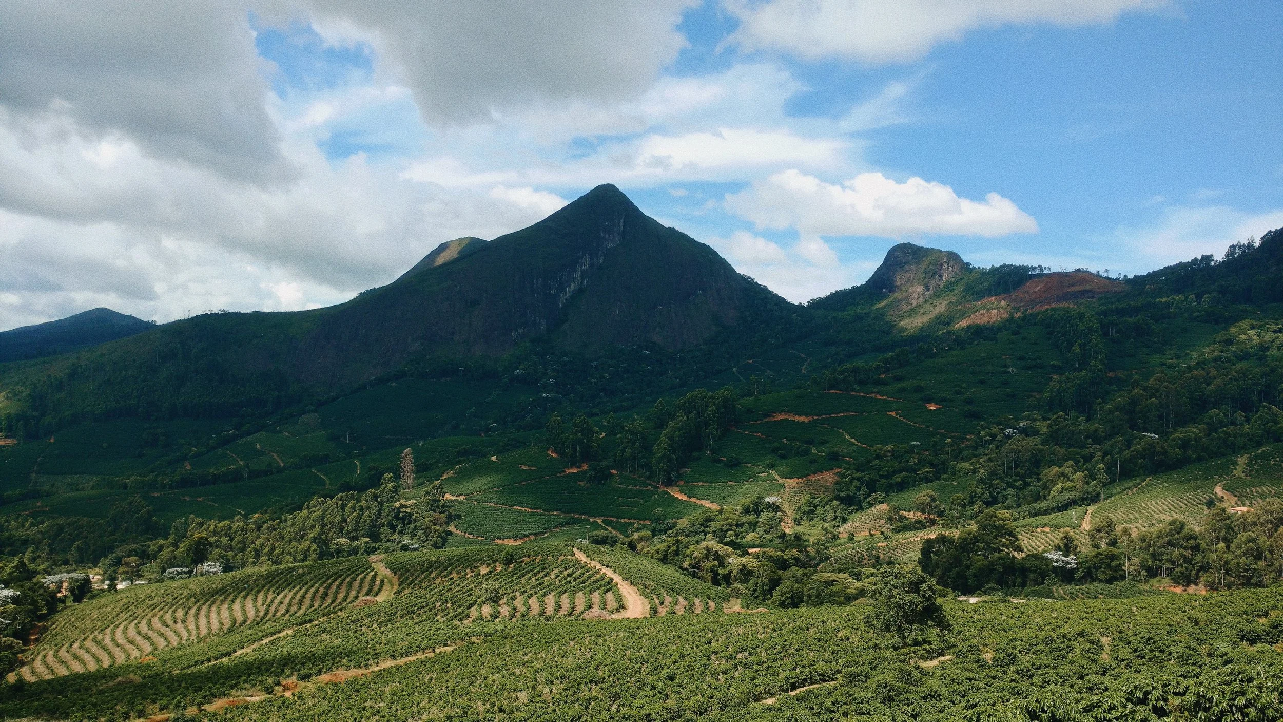 Lush green mountains with cultivated fields and rolling hills under a partly cloudy sky. Matas De Minas Fazendas Dutra