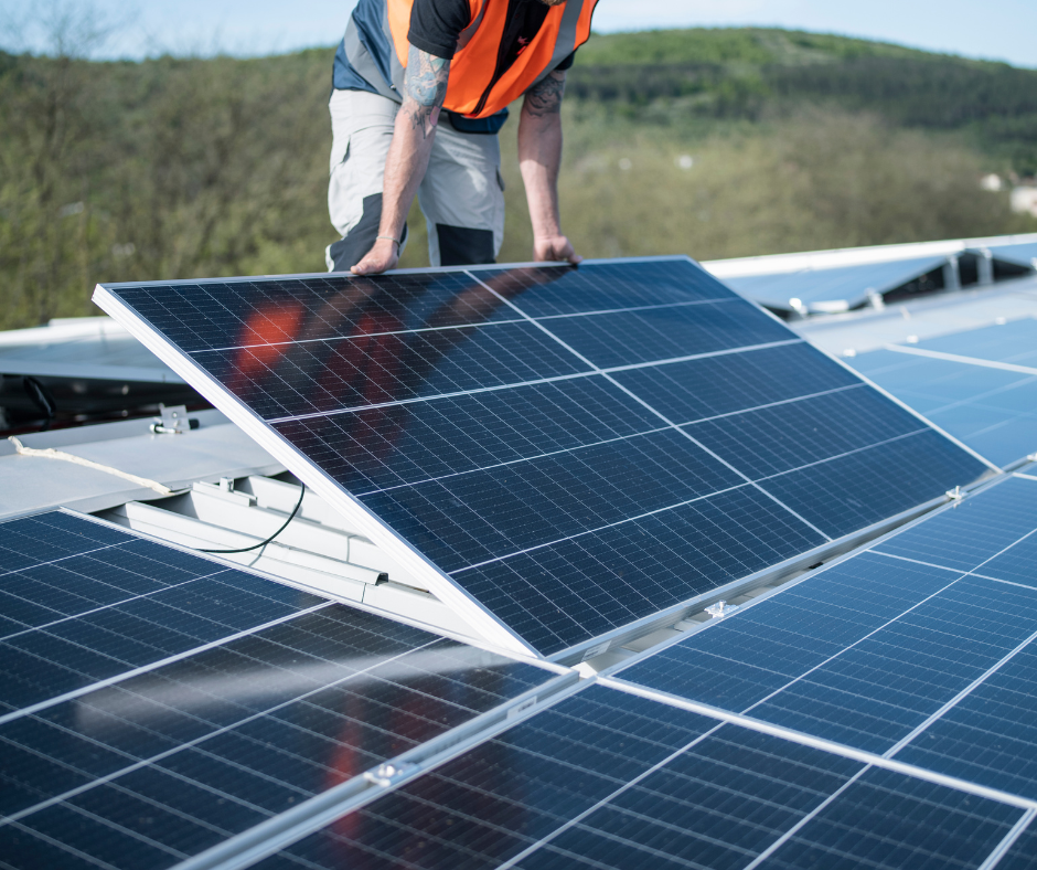 A worker installs solar panels on a rooftop, with green hills in the background.