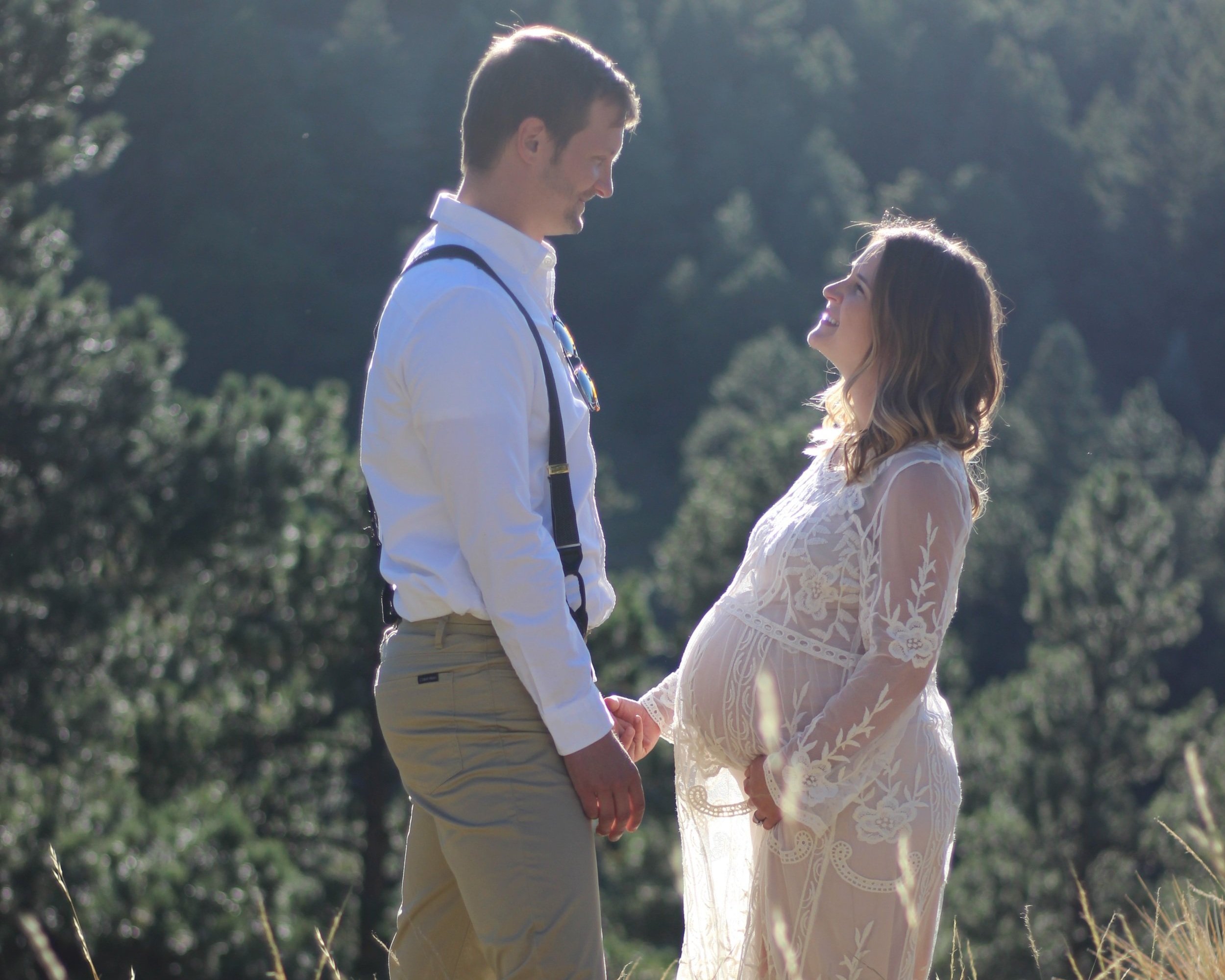 A couple standing outdoors in a scenic, wooded area, holding hands and smiling at each other, with the woman visibly pregnant.