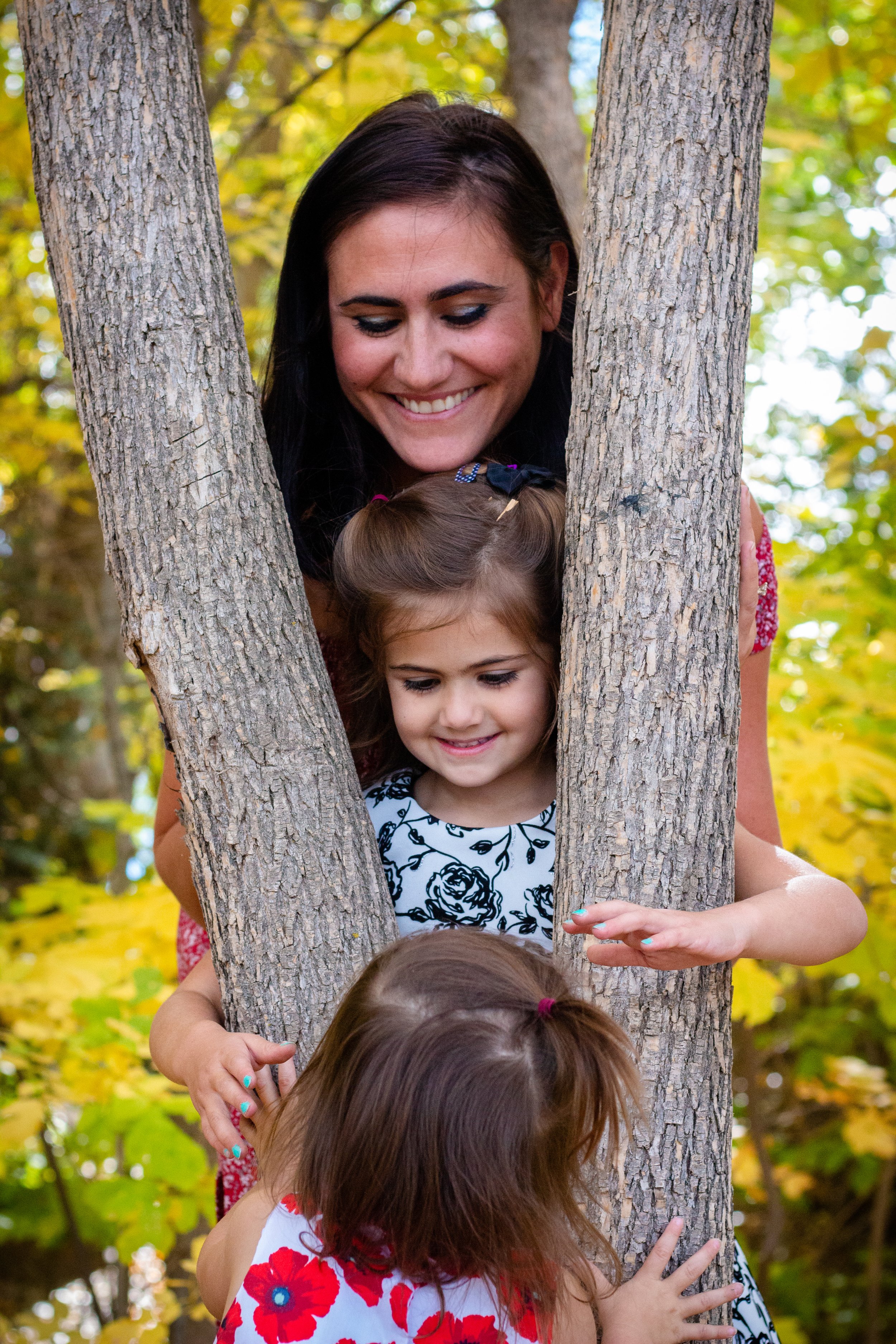 A woman smiling and hugging two young girls while playfully peeking through a tree in a wooded park area with yellow and green leaves.