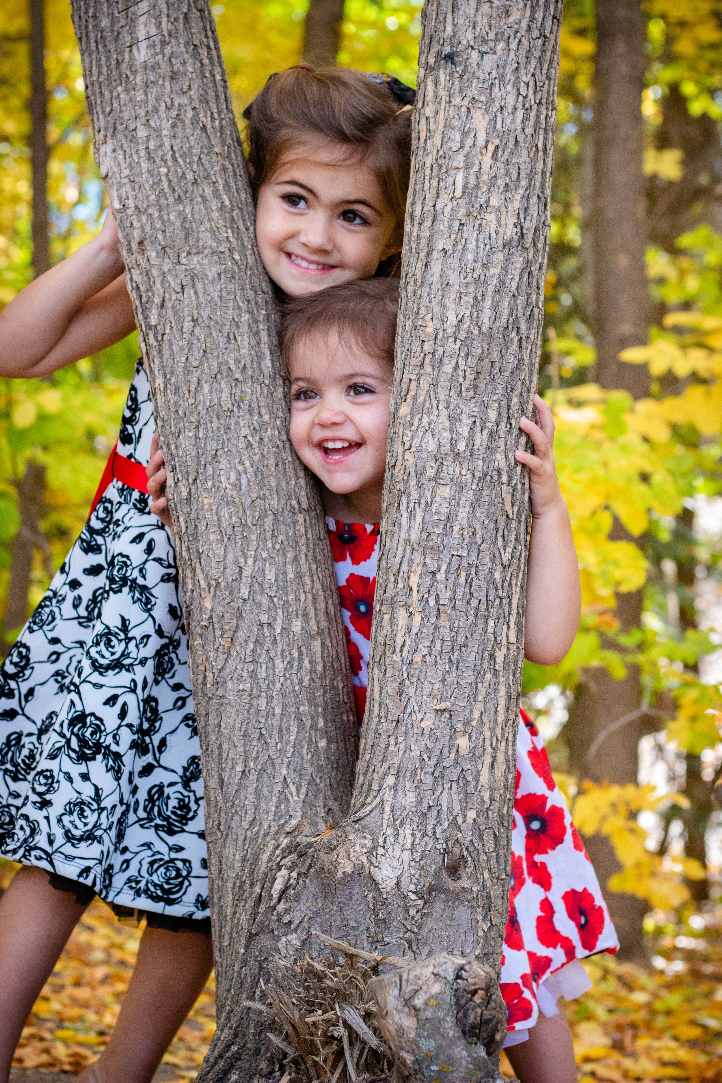 Two young girls peeking from behind a tree in a forest, smiling and playing together.