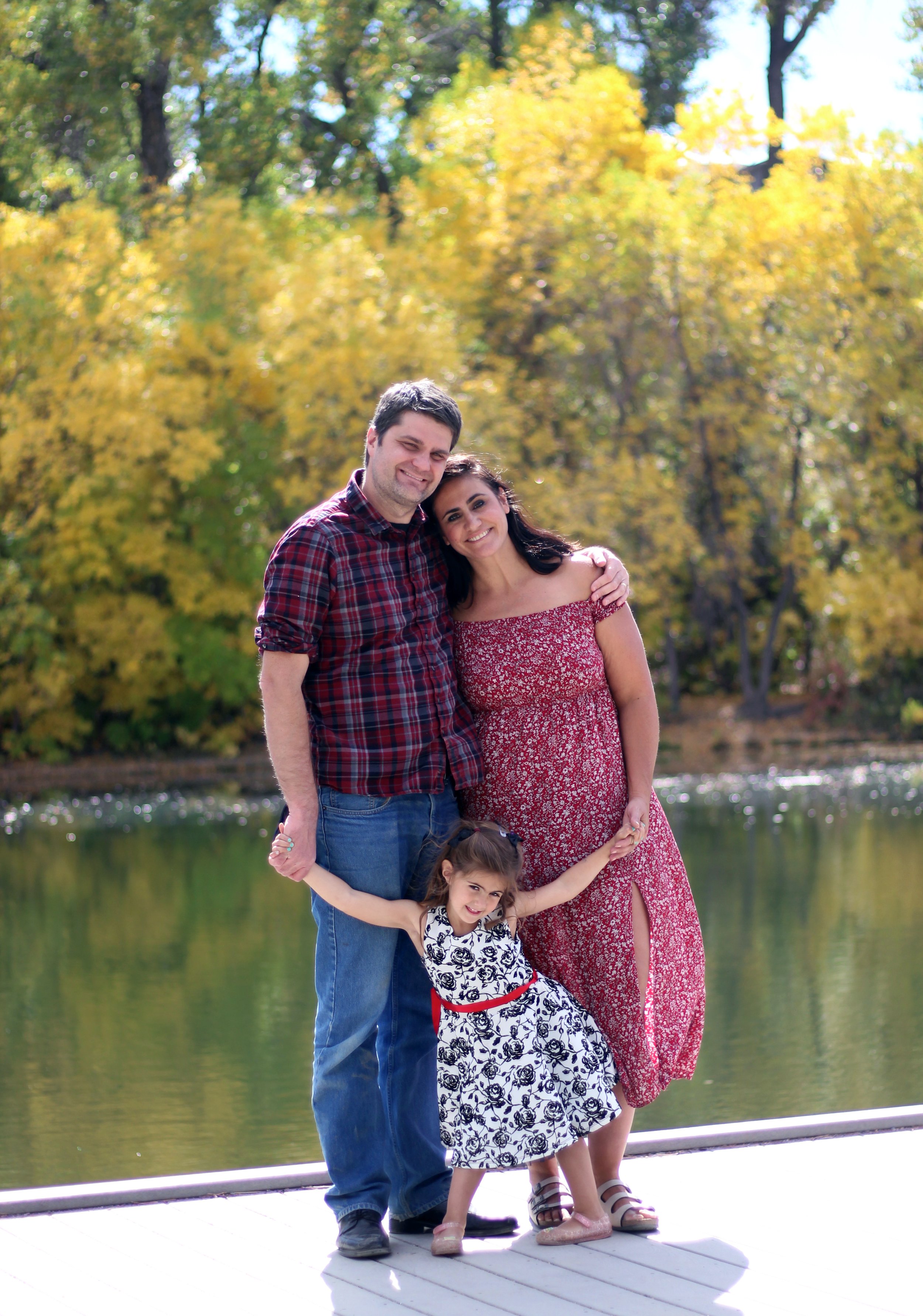 A family of three standing outdoors near a lake with trees in fall colors, smiling and embracing. The man is wearing a red plaid shirt and jeans, the woman is in a red dress with a small slit, and the young girl wears a black and white floral dress w
