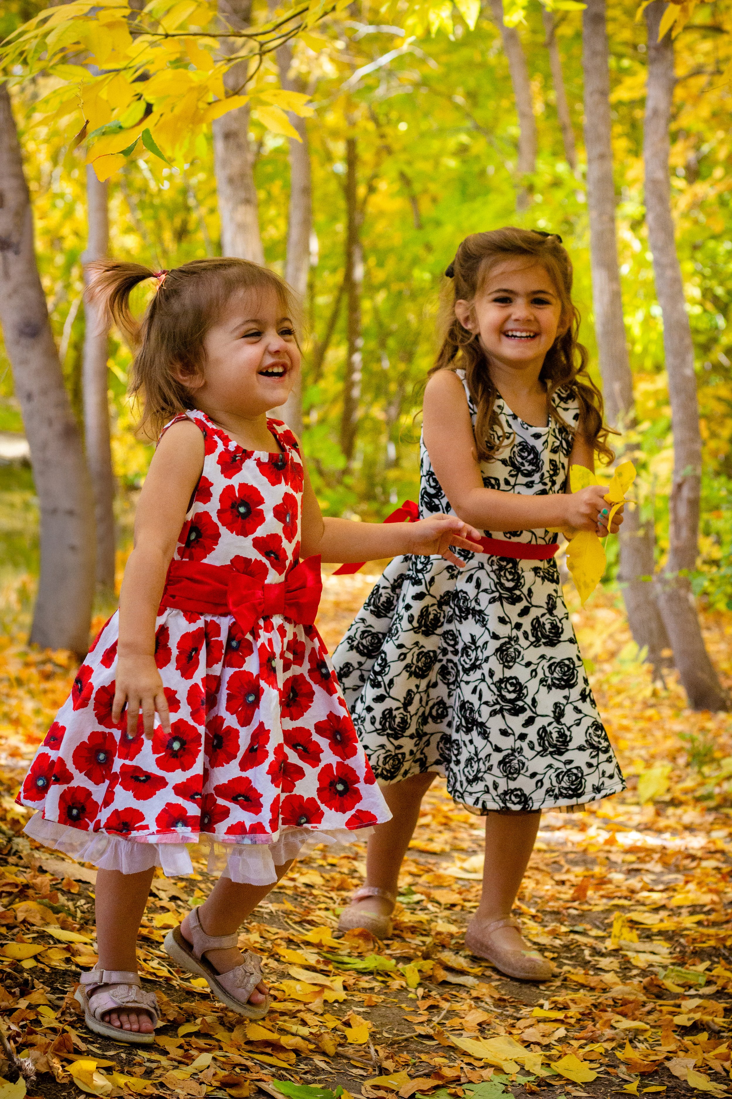 Two young girls walking in a forest during fall, smiling and laughing. One girl is wearing a red and white floral dress with a large red bow, and the other girl is wearing a black and white floral dress. Fallen leaves cover the ground and trees with