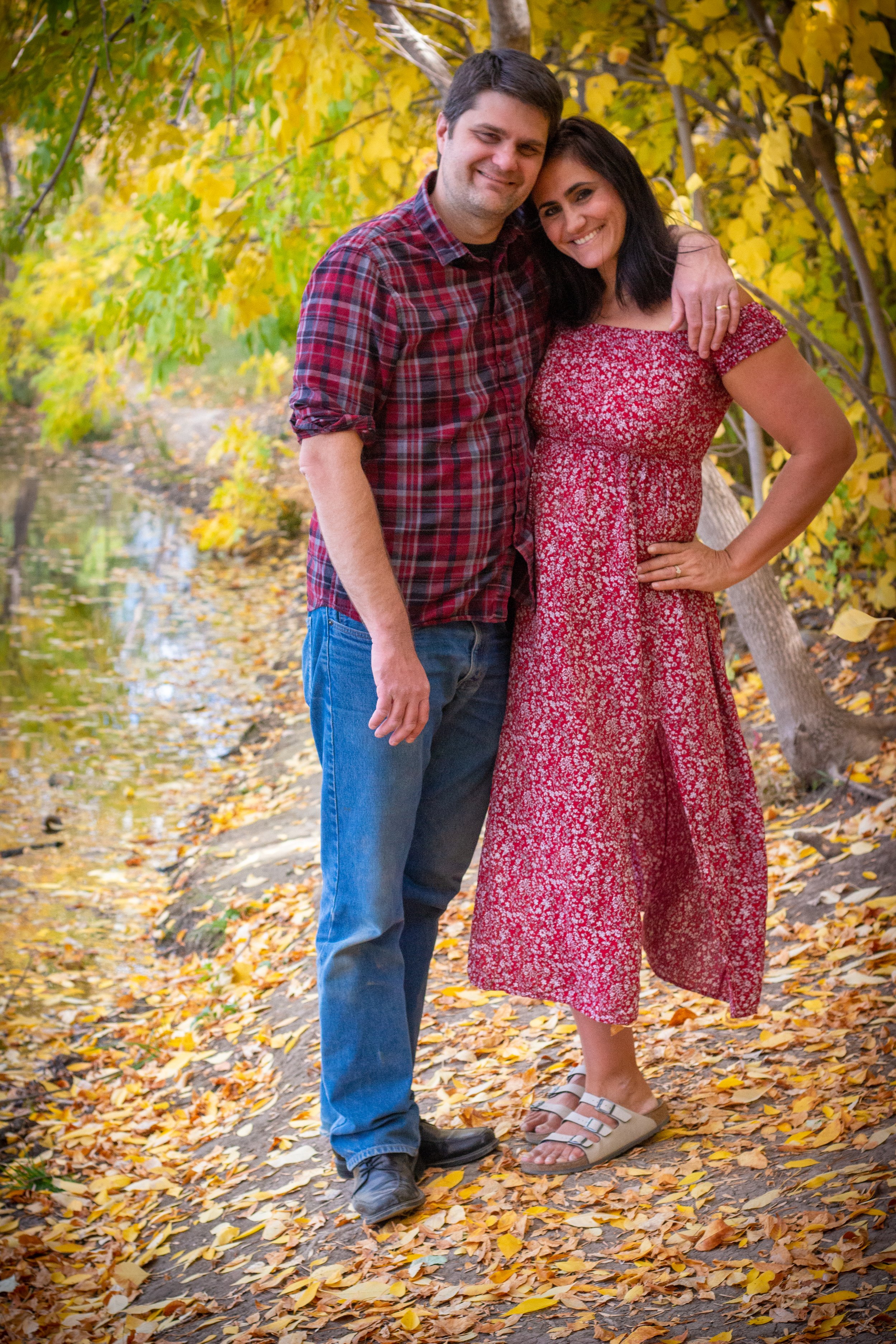 A smiling man and woman embrace outdoors on a fall day, surrounded by yellow and orange leaves and trees, standing on a leaf-covered trail near a body of water.