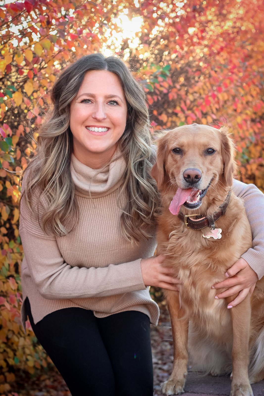 A woman with long, wavy, light brown hair smiling and sitting next to a happy golden retriever dog outdoors with colorful autumn leaves in the background.