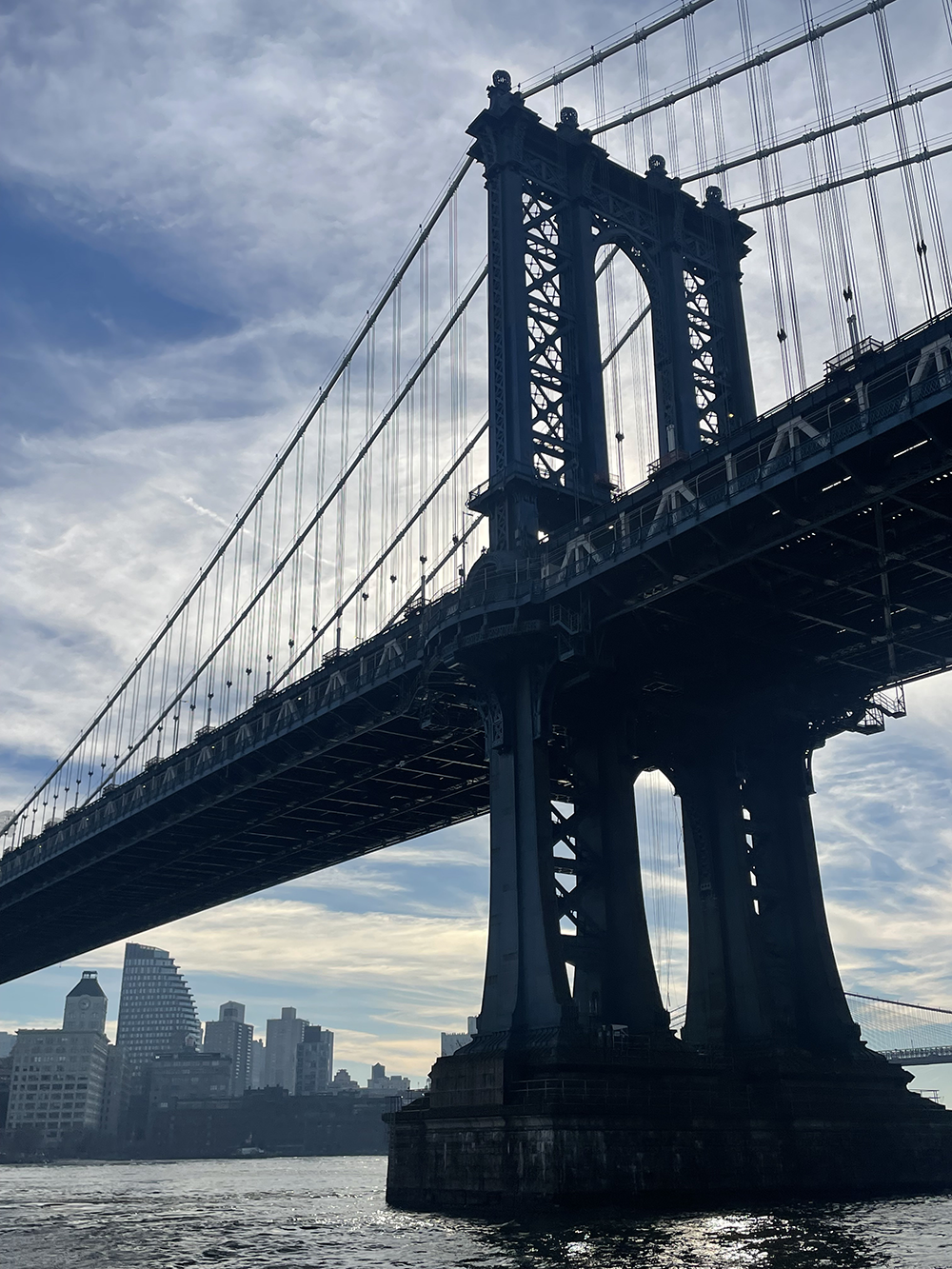 A view of the Brooklyn Bridge with its towers and suspension cables, with New York City skyline in the background and water in the foreground.