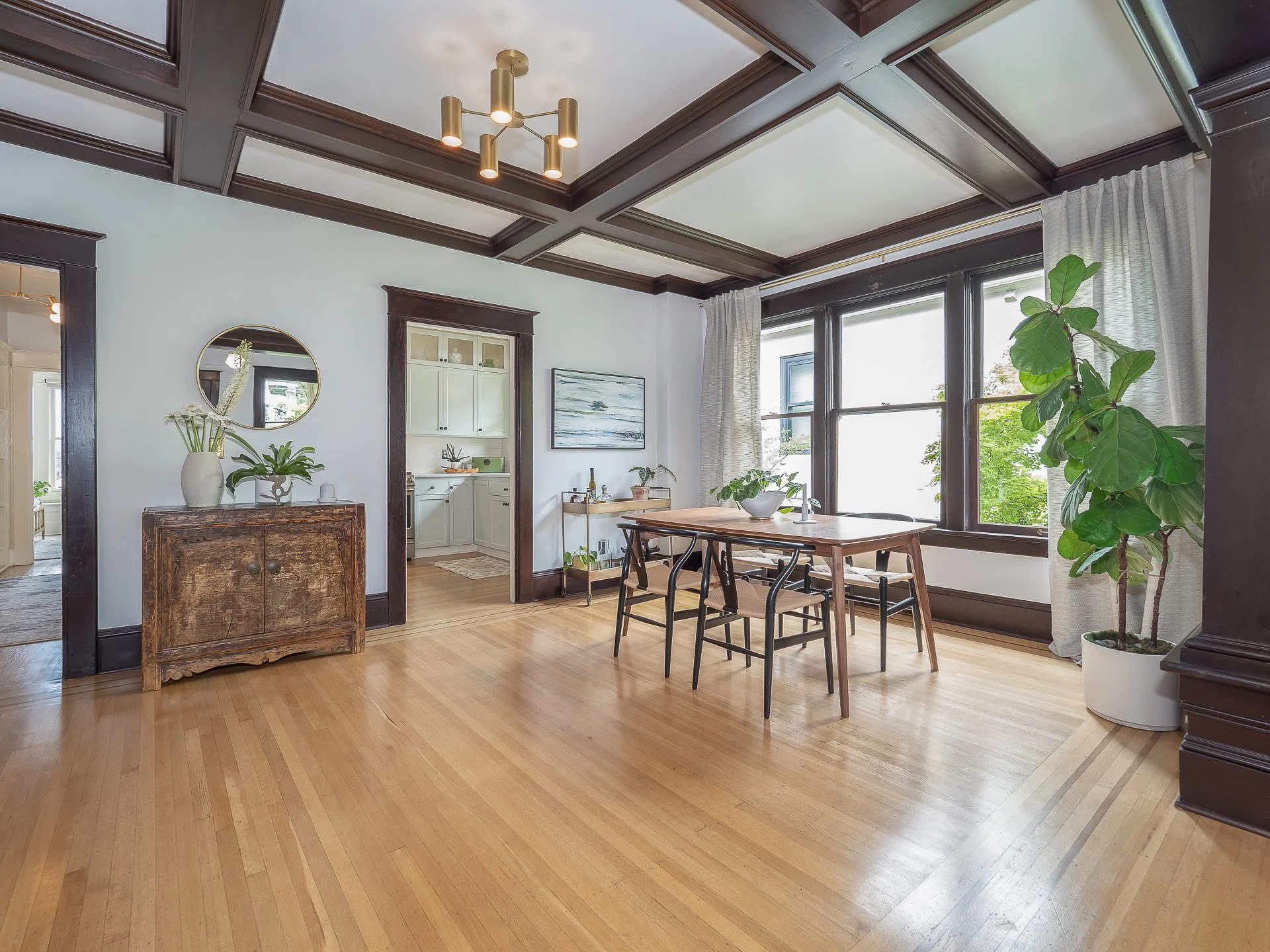 Bright dining room with hardwood floors, large windows with white curtains, wooden dining table, several black chairs, and large potted plant near window. Glass cabinet and wood console with decorative items are visible.