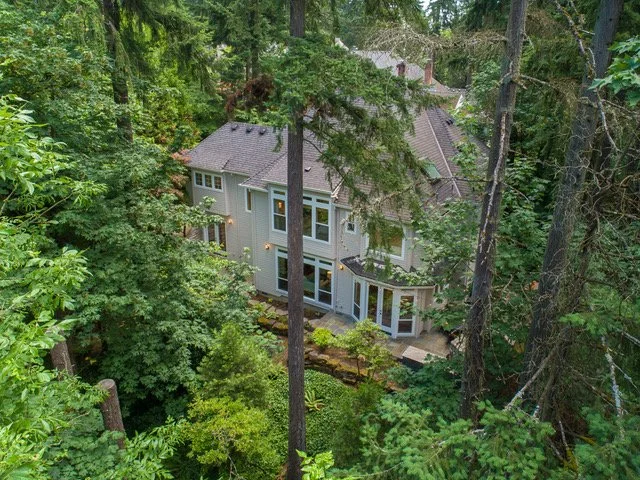 A large house surrounded by dense forest with tall trees and lush greenery, with visible windows and a deck area.