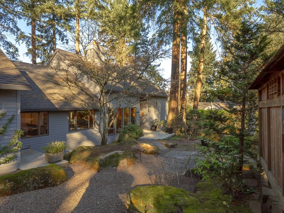 A cozy house surrounded by tall trees and a natural garden with rocks, potted plants, and a gravel pathway.