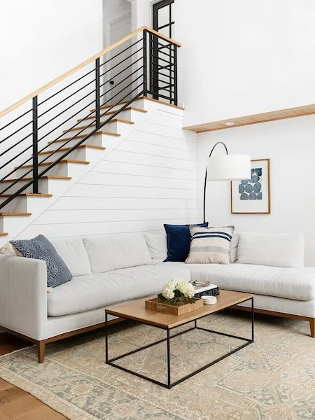 Living room with white sofa, throw pillows, a wooden coffee table, a floor lamp, artwork on the wall, and a staircase with black railing.