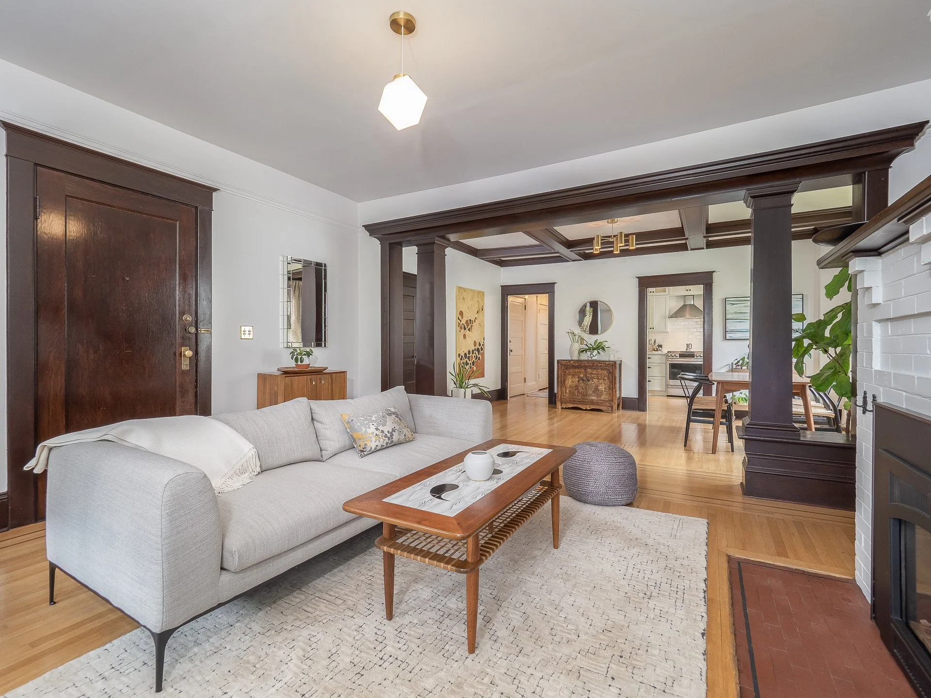 Living room with white sofa, wooden coffee table, gray pouf, white brick fireplace, and dark wood trim and beams.
