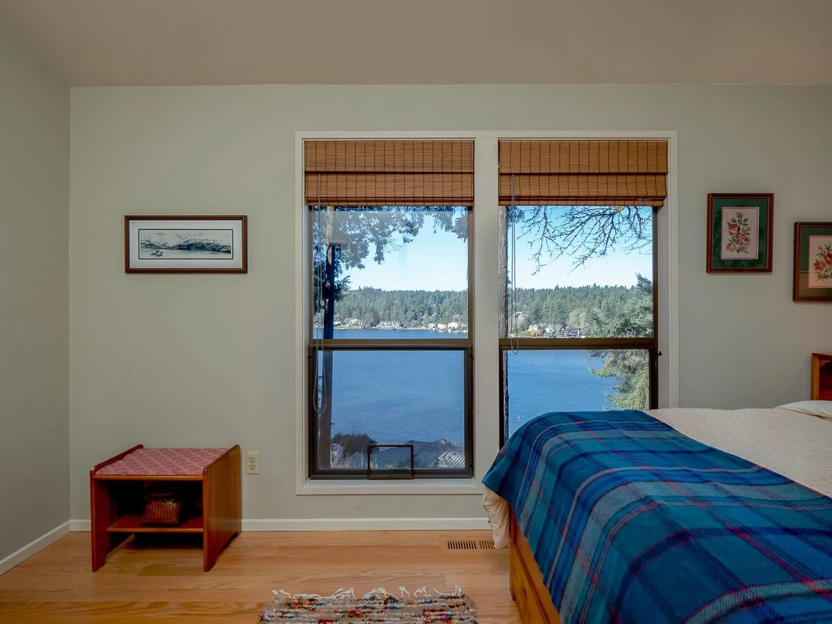 Bedroom with two windows overlooking a lake and trees, partially covered with bamboo blinds, bed with blue plaid blanket, small wooden side table, wall art, and hardwood floor.