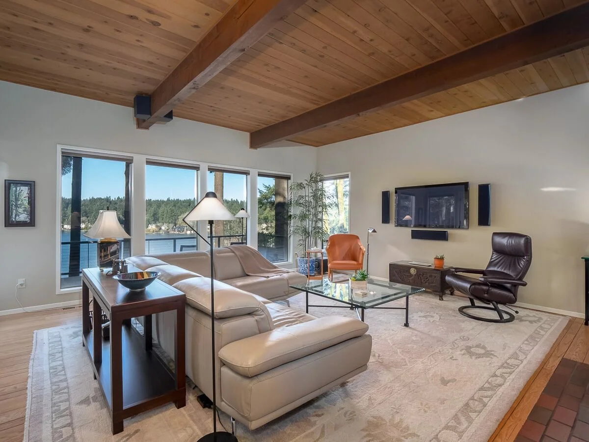 Modern living room with beige sectional sofa, glass coffee table, leather swivel chair, window views of water and trees, wooden ceiling with exposed beams, flat-screen TV, and decorative plants.