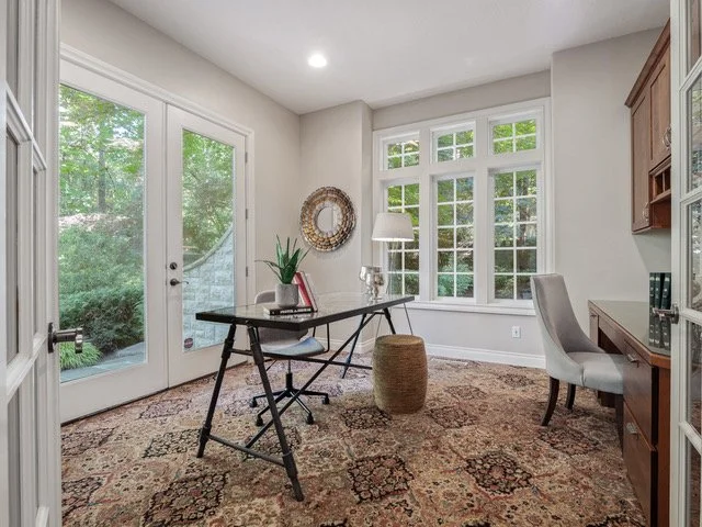 Elegant home office with large window overlooking greenery, glass door, desk with a chair, a plant, mirror, and neutral decor.