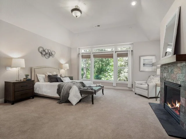 Spacious bedroom with a large window, beige carpet, bed with white bedding and gray accents, a fireplace, and modern decor.