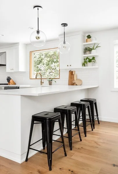 Modern white kitchen with a breakfast bar, black metal stools, pendant lights, wood floors, and open shelves with plants.