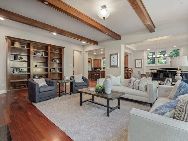 Living room with white sofa, armchairs, wooden coffee table with decorative items, built-in wooden bookshelves, hardwood floors, and exposed ceiling beams.