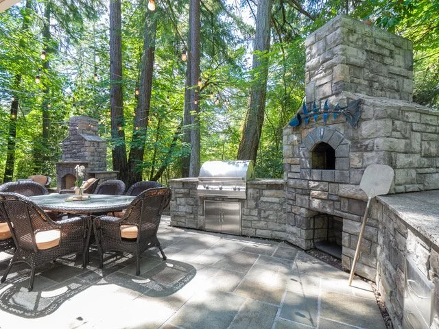 Outdoor stone patio area with a dining table, chairs, a built-in grill, and a fireplace, surrounded by tall trees and string lights.