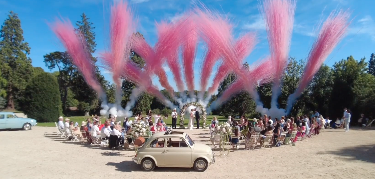 Cérémonie de mariage en plein air avec des invités assis, des fleurs et un portique. Feux d'artifice colorés rose dans le ciel, voitures anciennes autour de l'événement.