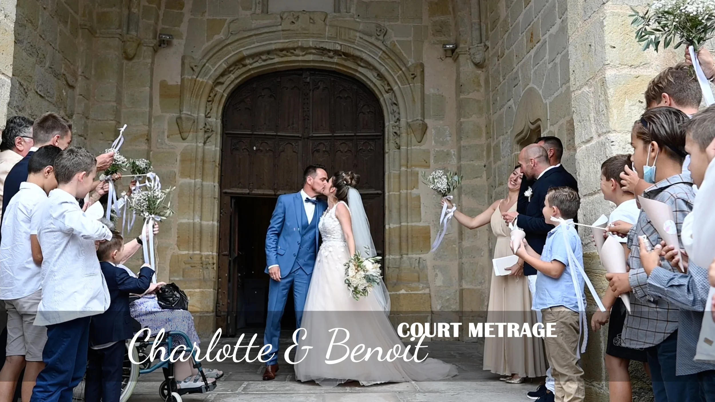 Un couple de mariés, Charlotte et Benoît, s'embrassent devant une porte en pierre d'une vieille église, entourés d'invités qui leur lancent des confettis et tiennent des bouquets ou des cônes à confettis, dans une célébration de mariage en extérieur.