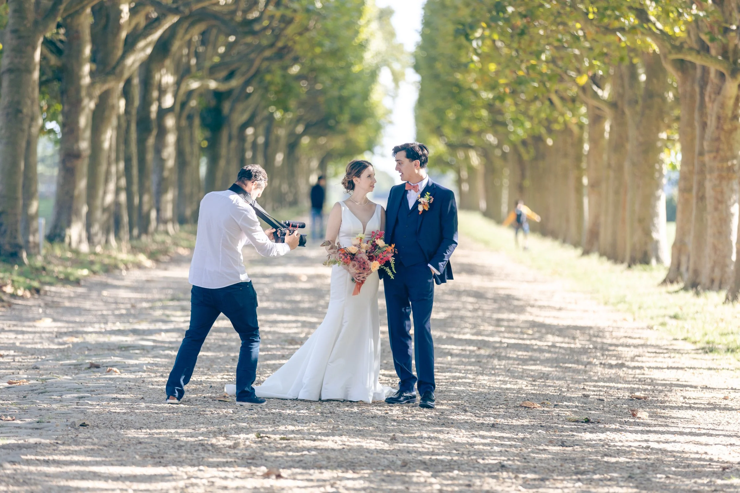 Un couple de mariés en costume noir et robe blanche, se tenant dans une allée de arbres durant un mariage, terrasse un bouquet de fleurs colorées, pendant qu'un caméraman filme la scène, en plein air par une journée ensoleillée.