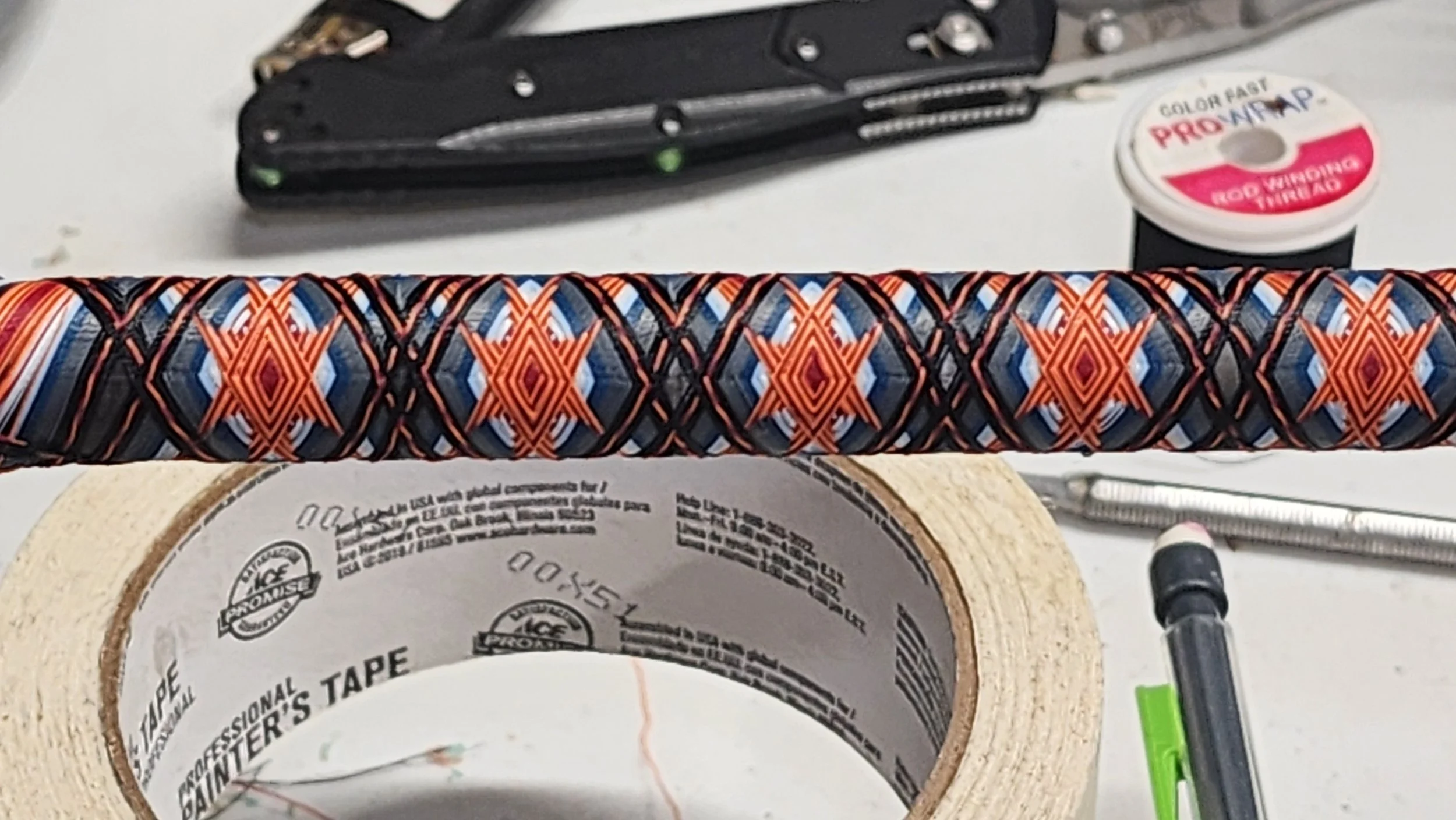 Close-up of a golf club grip with a patterned orange, black, and gray design on a white work surface. Surrounding objects include a roll of painter's tape, a black pen with a green clip, a mechanical pencil, a black multi-tool, and a spool of Pro Wrap. The backdrop is slightly out of focus.