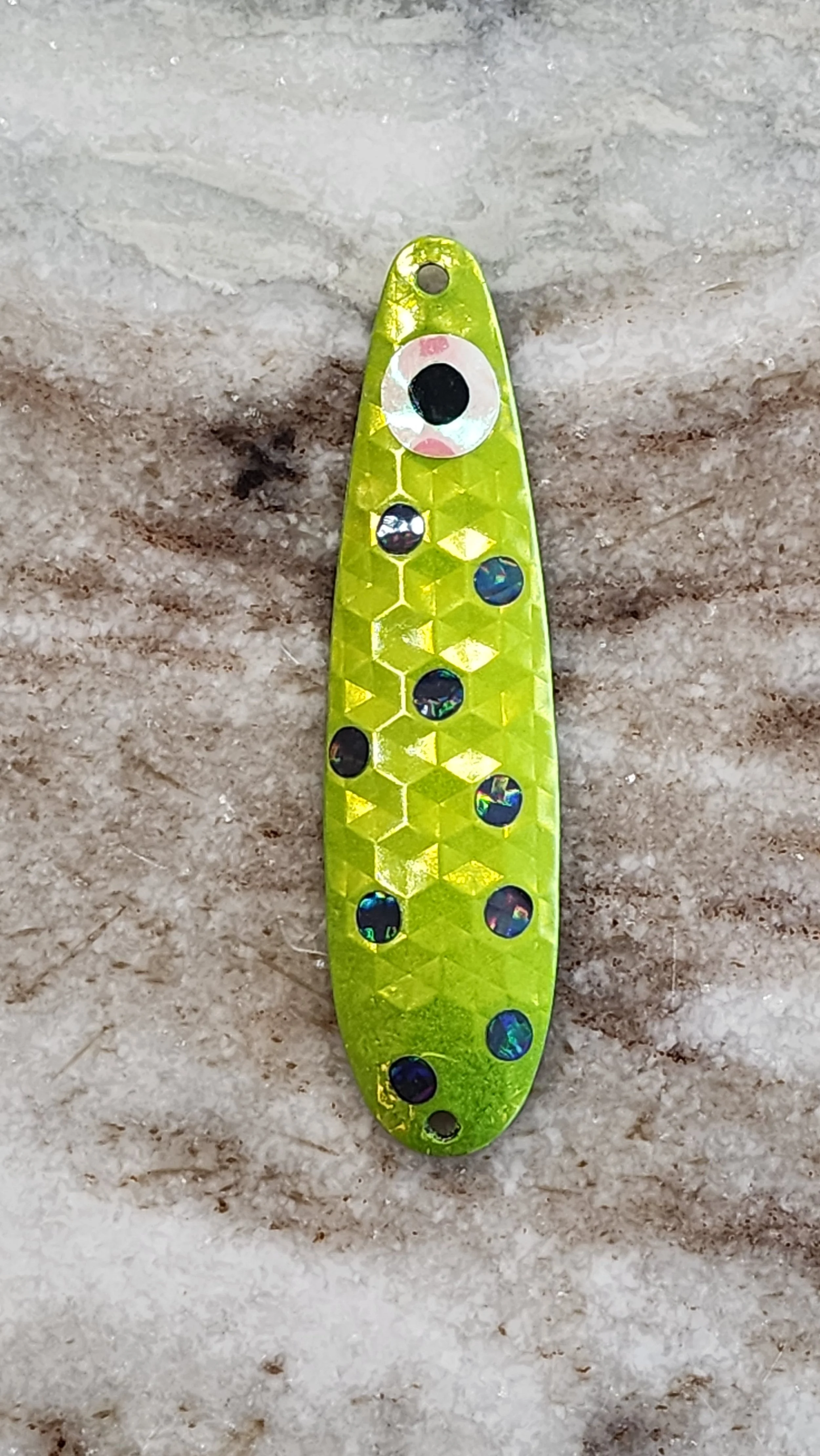 A yellow fishing lure with a honeycomb pattern and holographic circular accents, positioned against a sandy background.