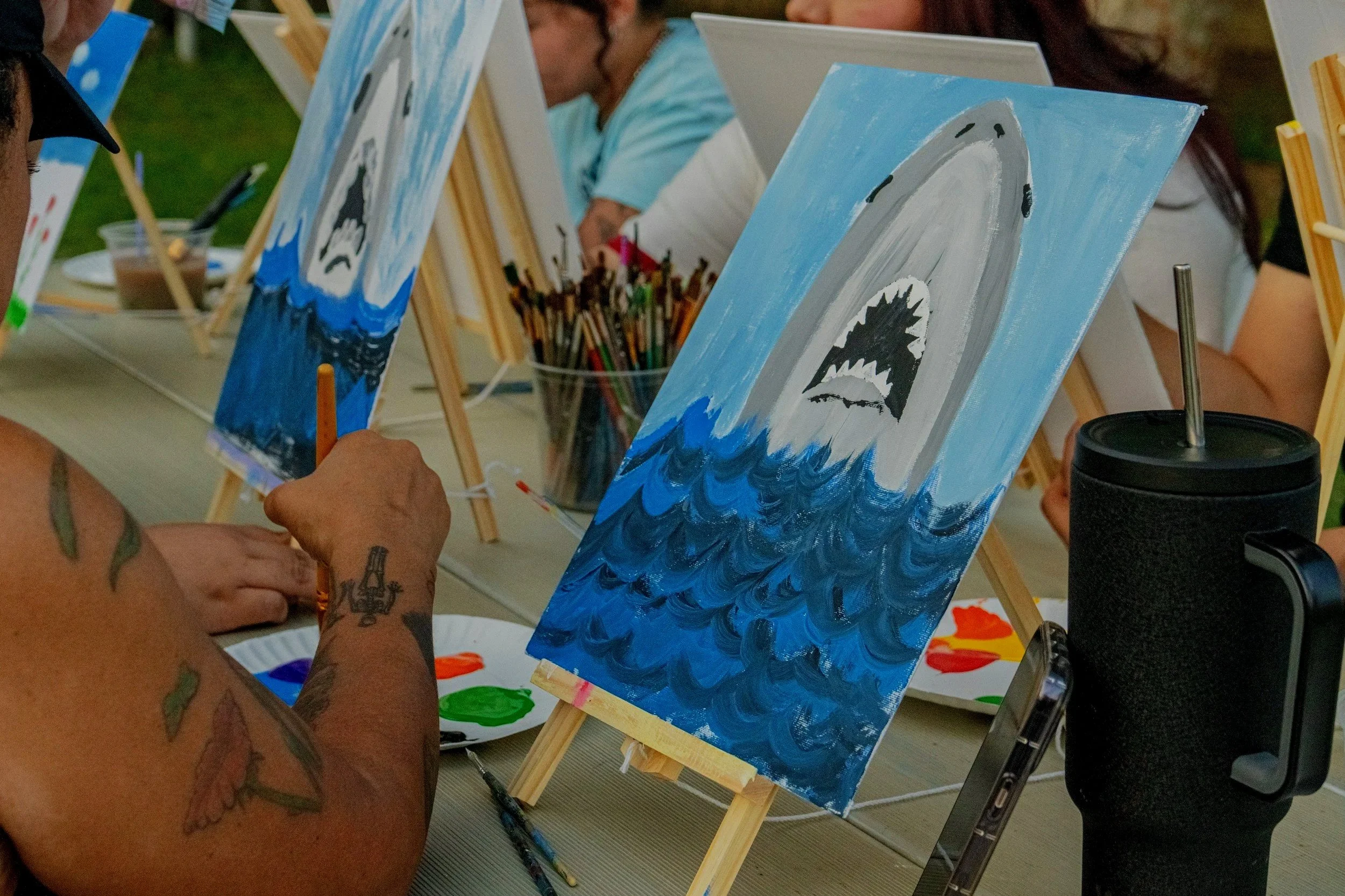 People painting a large shark fin emerging from the water on canvases at an outdoor painting event.