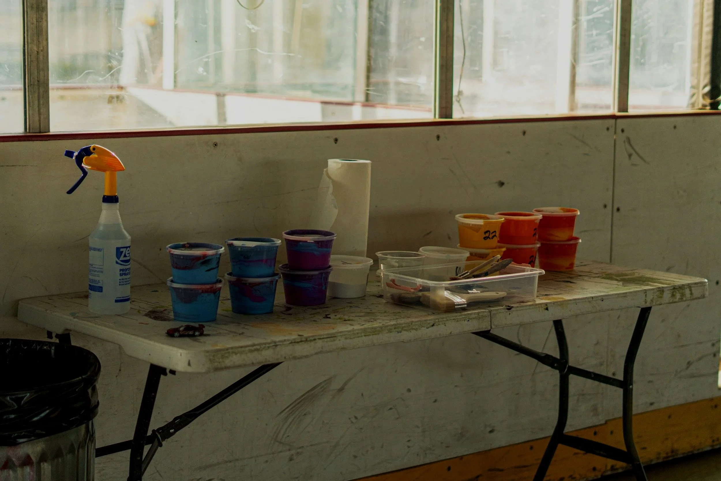 A weathered table with colorful paint pots, brushes, paper towels, and a spray bottle, set against a wall beneath a large window.