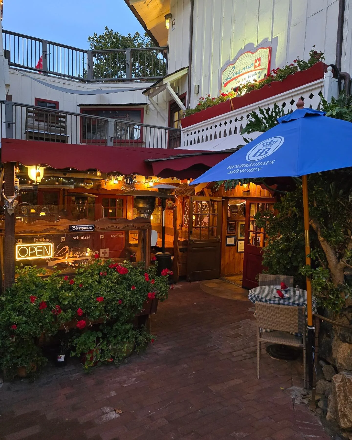 Outdoor seating area of a restaurant or cafe with a blue umbrella, a small checkered table, and potted plants, illuminated by warm outdoor lighting.