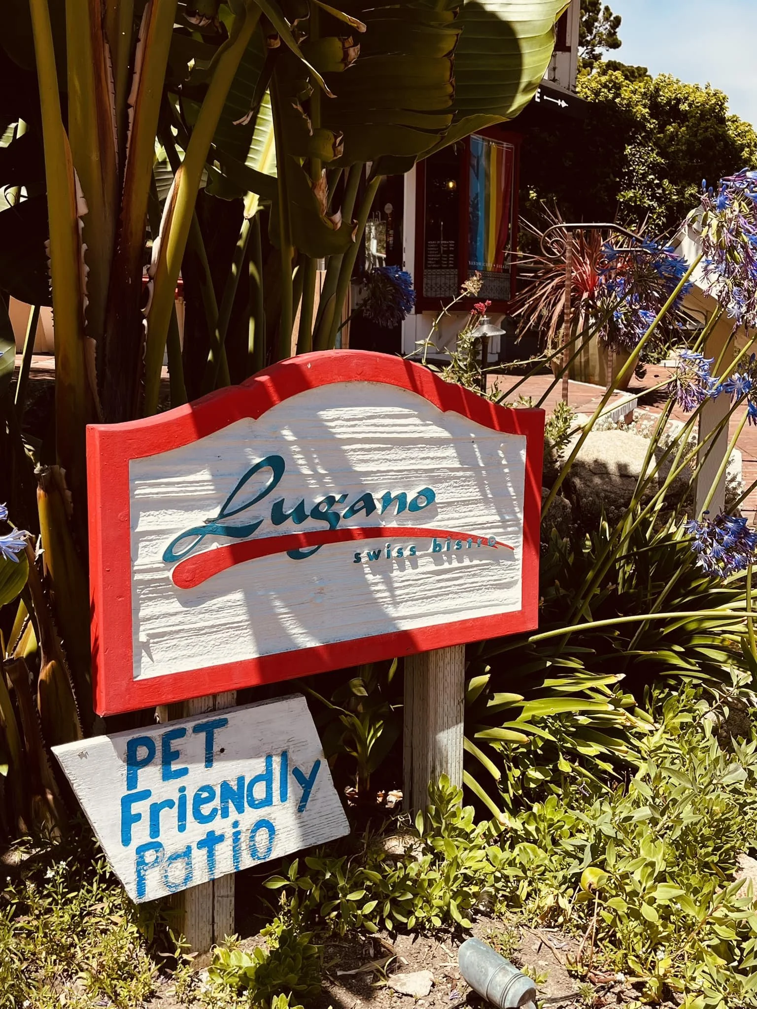 Sign for Lugano Swiss Bistro with a smaller sign below indicating pet-friendly patio, surrounded by tropical plants and flowers.