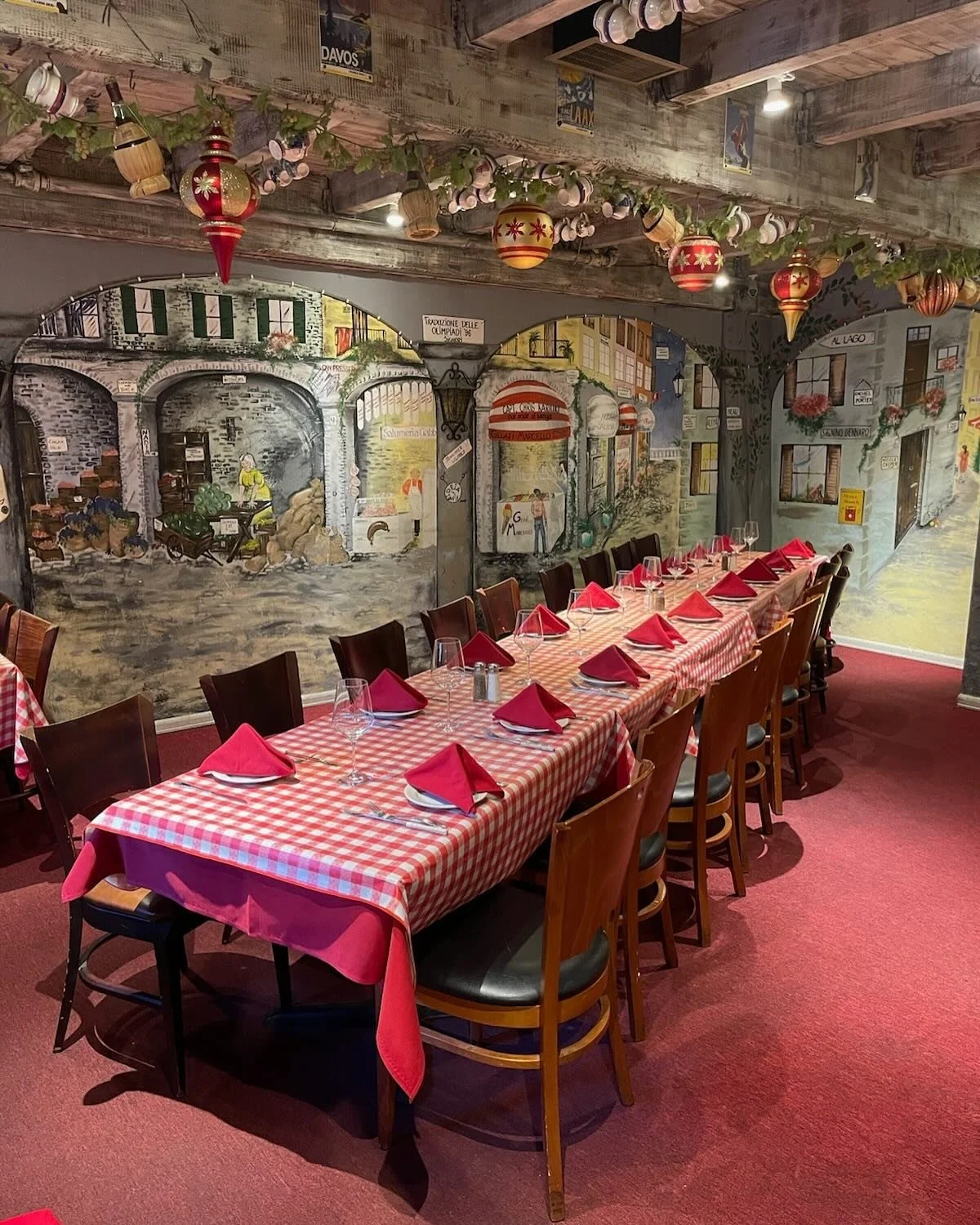A long dining table with a red and white checkered tablecloth, set with wine glasses, silverware, plates, and red cloth napkins in a restaurant with muraled walls and hanging holiday decorations.