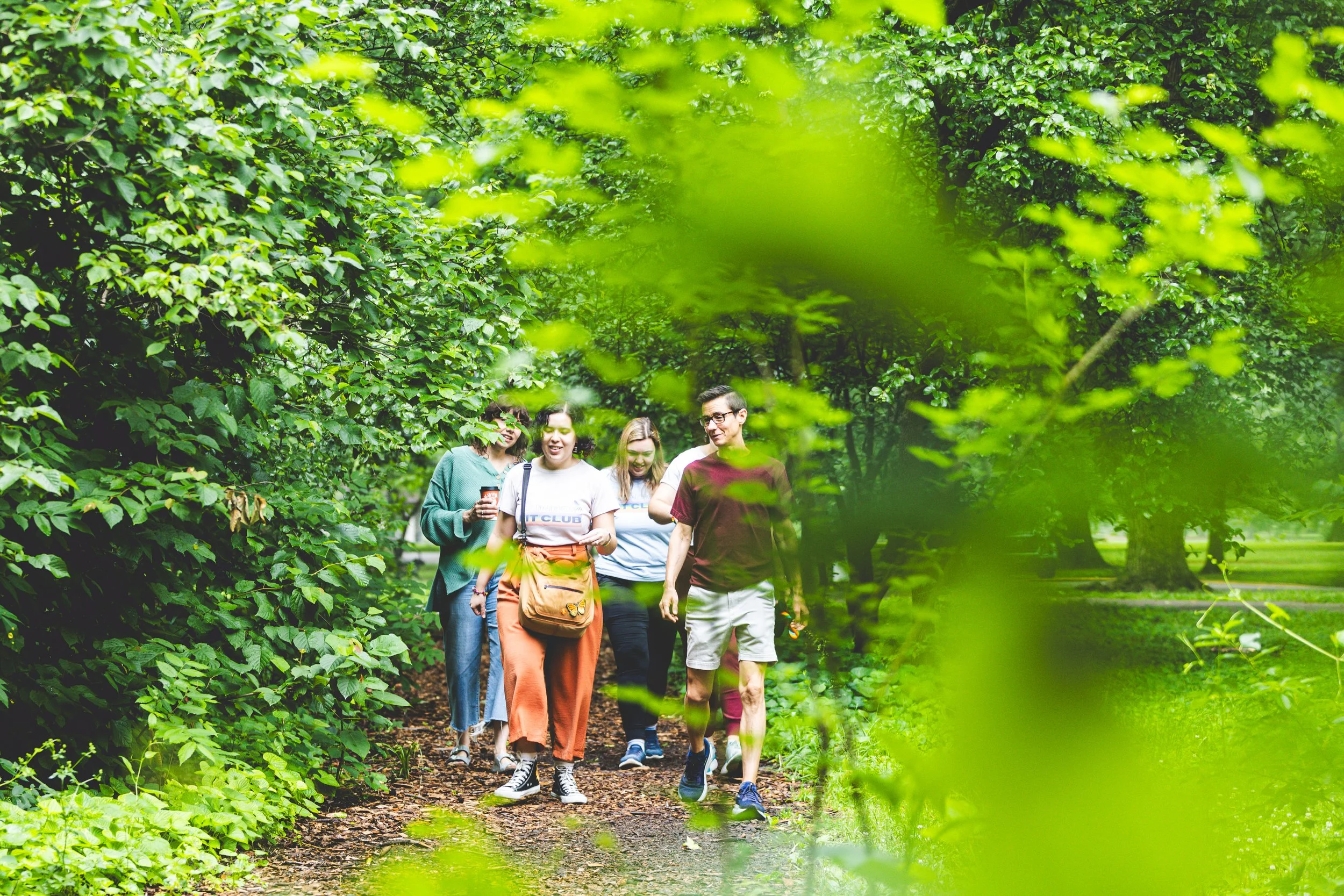 A group of five friends walking through a lush green forest trail while smiling and enjoying each other's company.