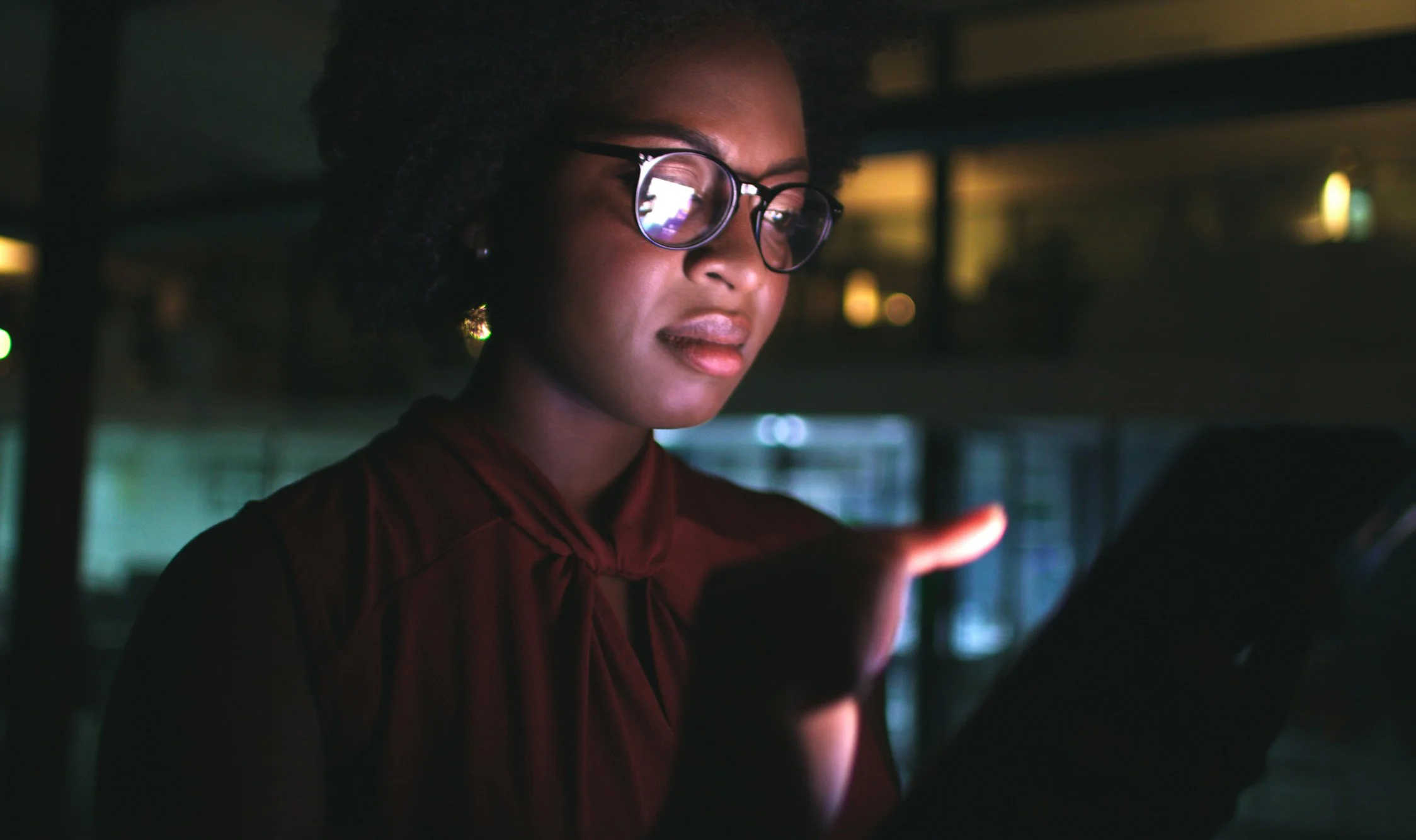 A woman wearing glasses looks at her phone in a dimly lit environment.