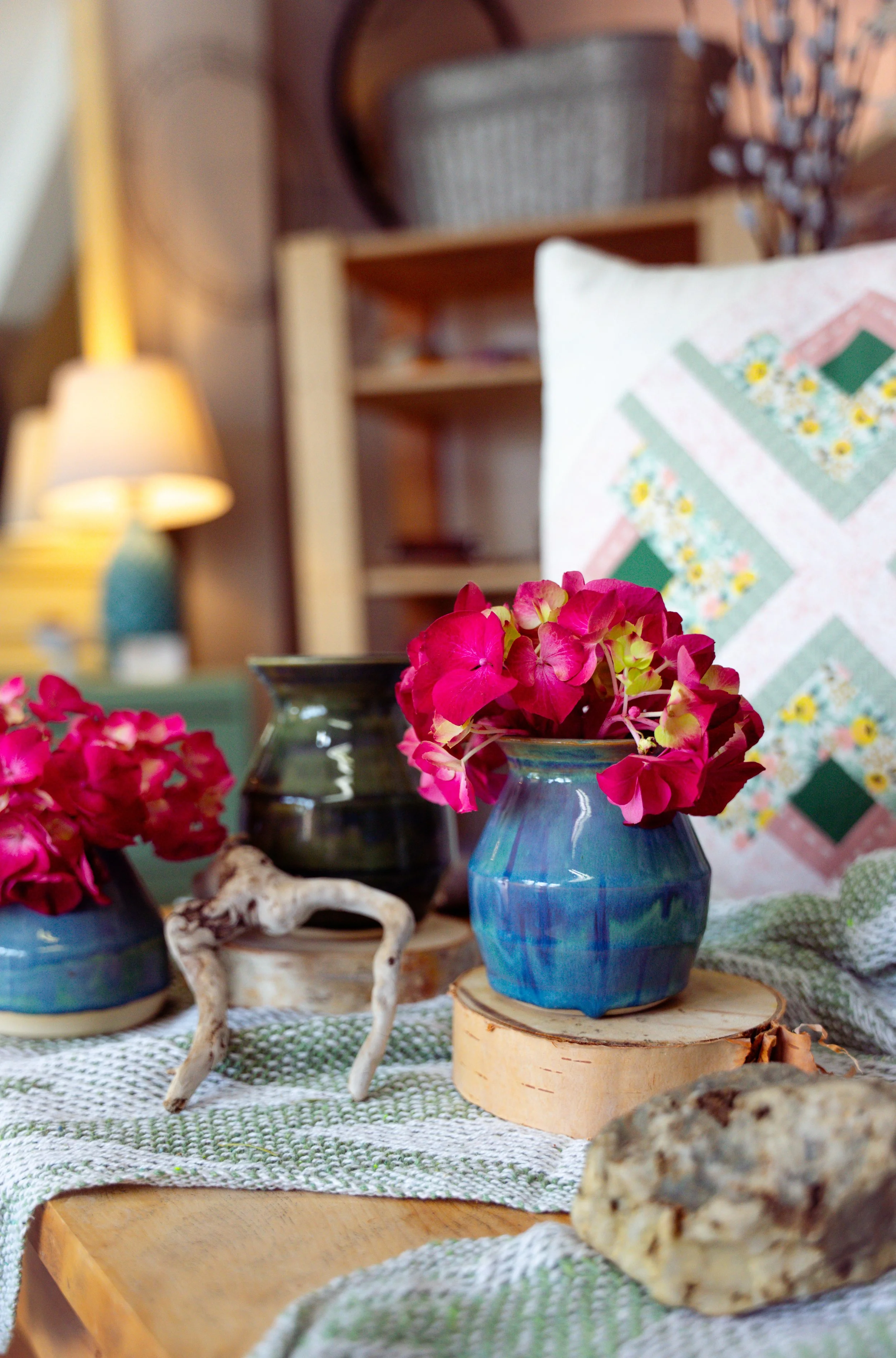 Decorative indoor scene with pink flowers in a blue vase, a green vase, and a dark vase on a table with a green and white cloth, a wooden slice, and a rock, with a quilted pillow and bookshelf in the background.