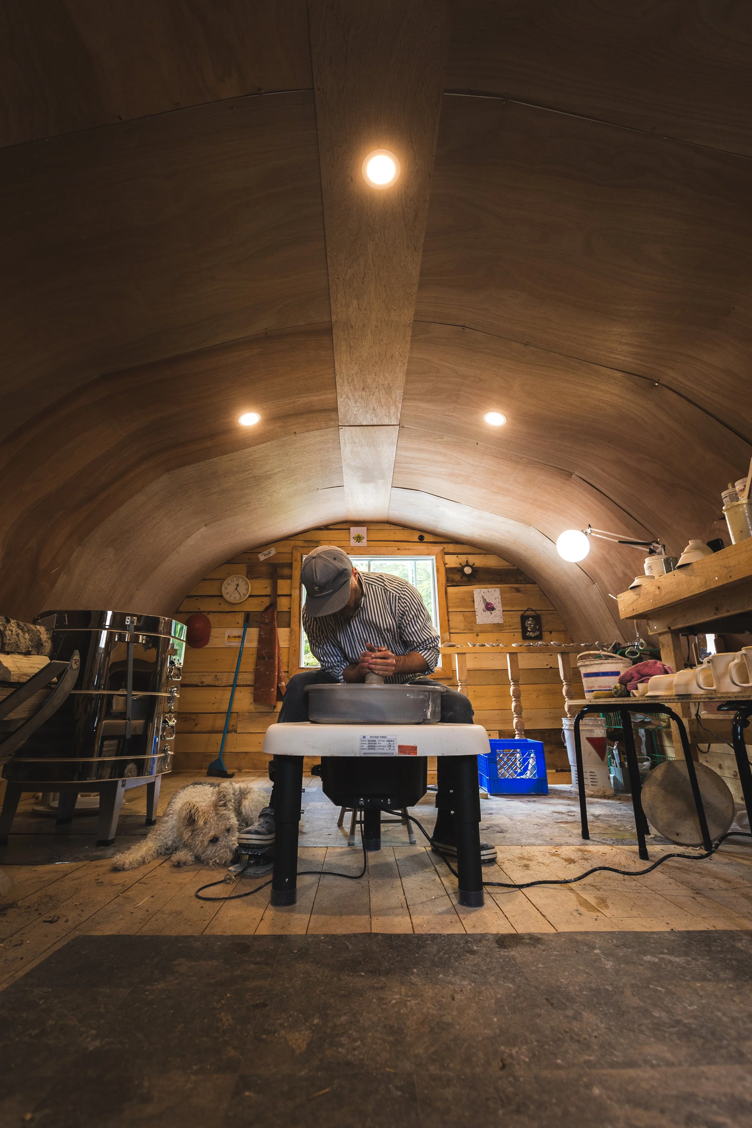 A person with a cap and striped shirt carving wood on a workbench inside a wooden arched room with various tools and decorations, a dog lying on the floor nearby, and warm lighting from ceiling bulbs.