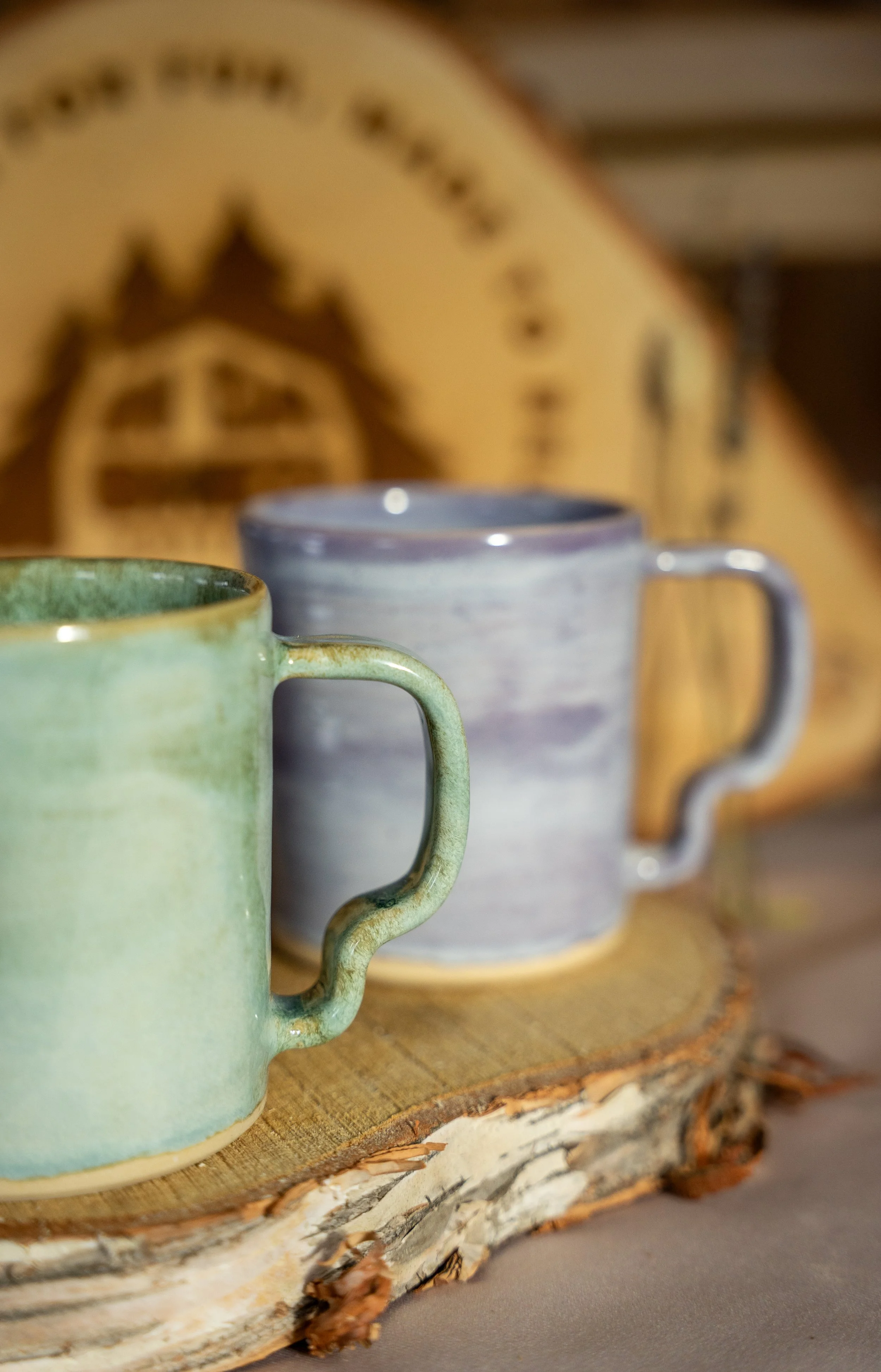 Two ceramic mugs, one green and one gray with purple streaks, placed on a wooden slab with bark edges, against a blurred background with a clock.
