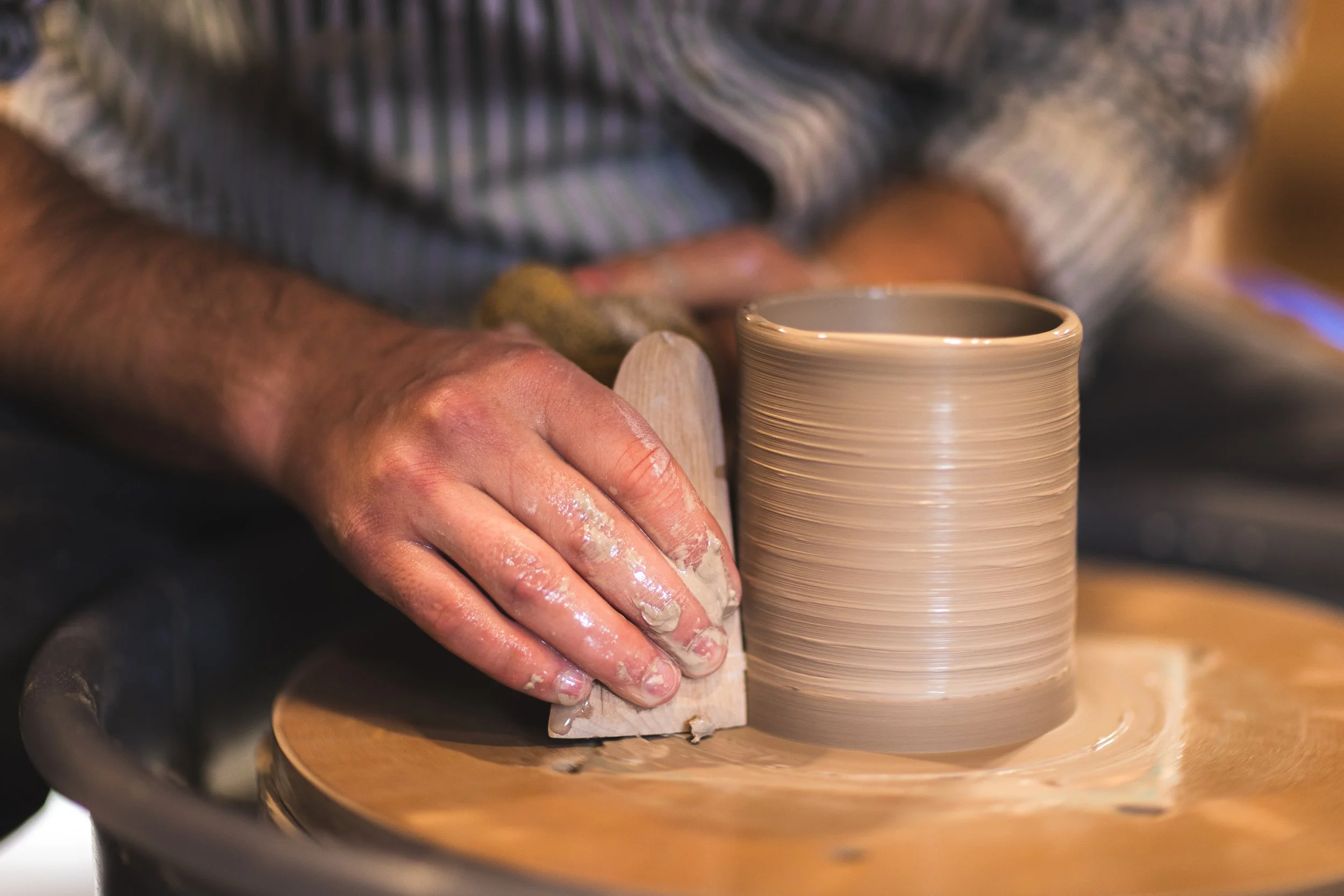 A person is shaping a ceramic cup on a pottery wheel, with clay on their hands and spinning wheel.