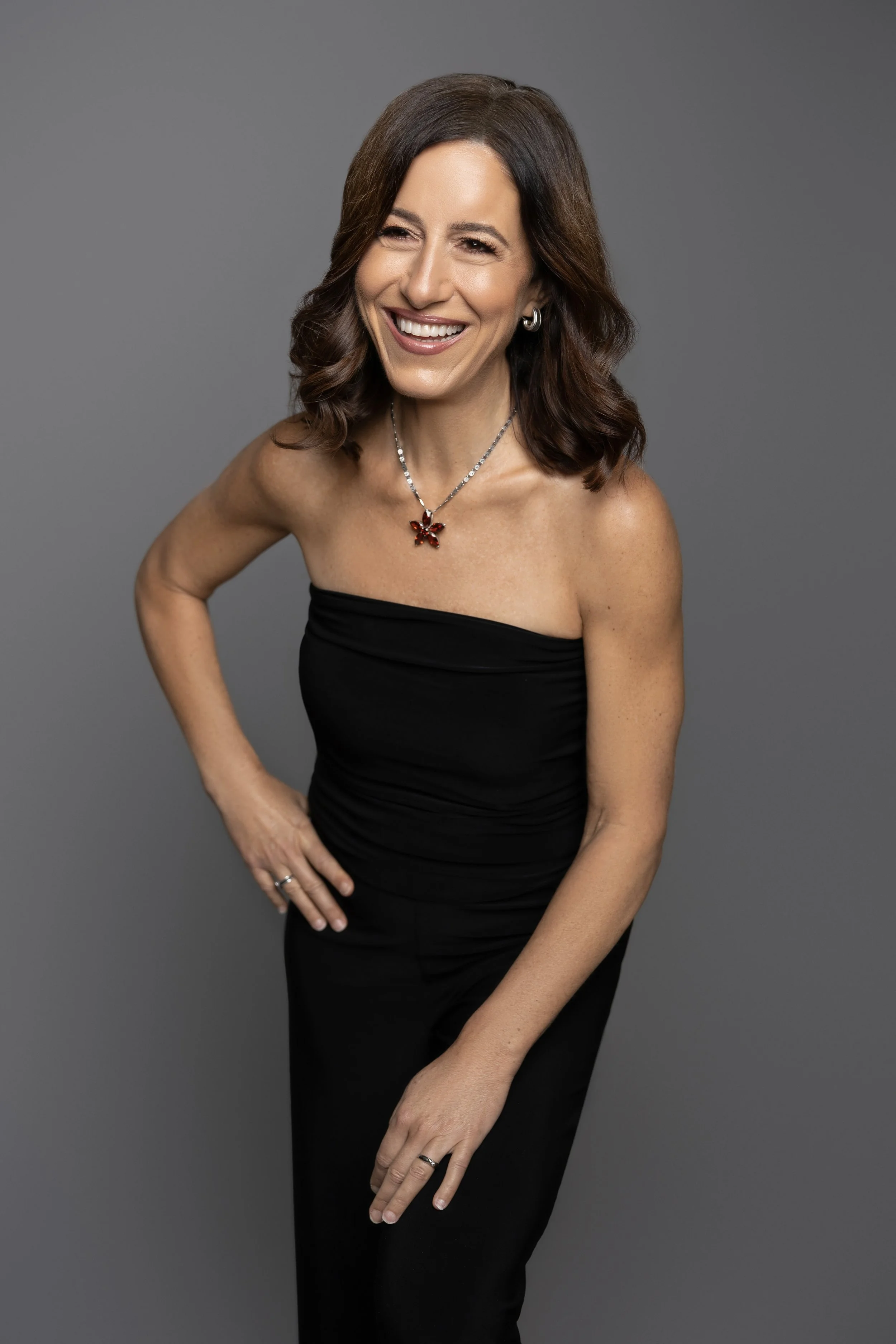 A woman with shoulder-length brown hair, wearing a strapless black dress, smiling and winking, standing against a gray background.
