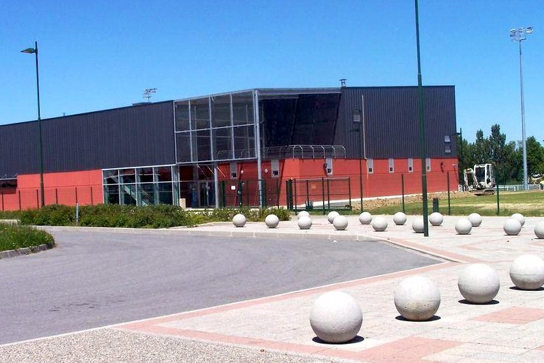 Un bâtiment sportif moderne avec un extérieur en métal noir et une partie rouge, entouré d'une allée pavée et de bornes rondes en béton.