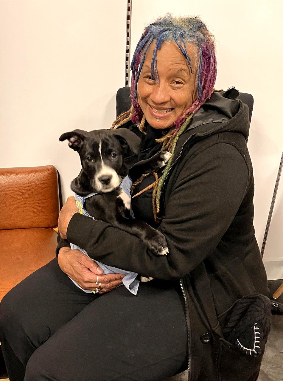 A woman with multicolored dreadlocks holding a small black and white therapy dog, sitting on a brown bench with white walls in the background.