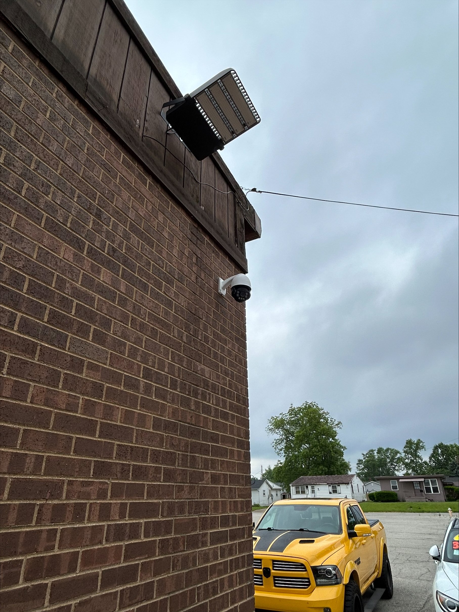A security camera is mounted on the brick wall of a building, with a large floodlight above it. In the background, a yellow pickup truck and other residential buildings are visible on a cloudy day.