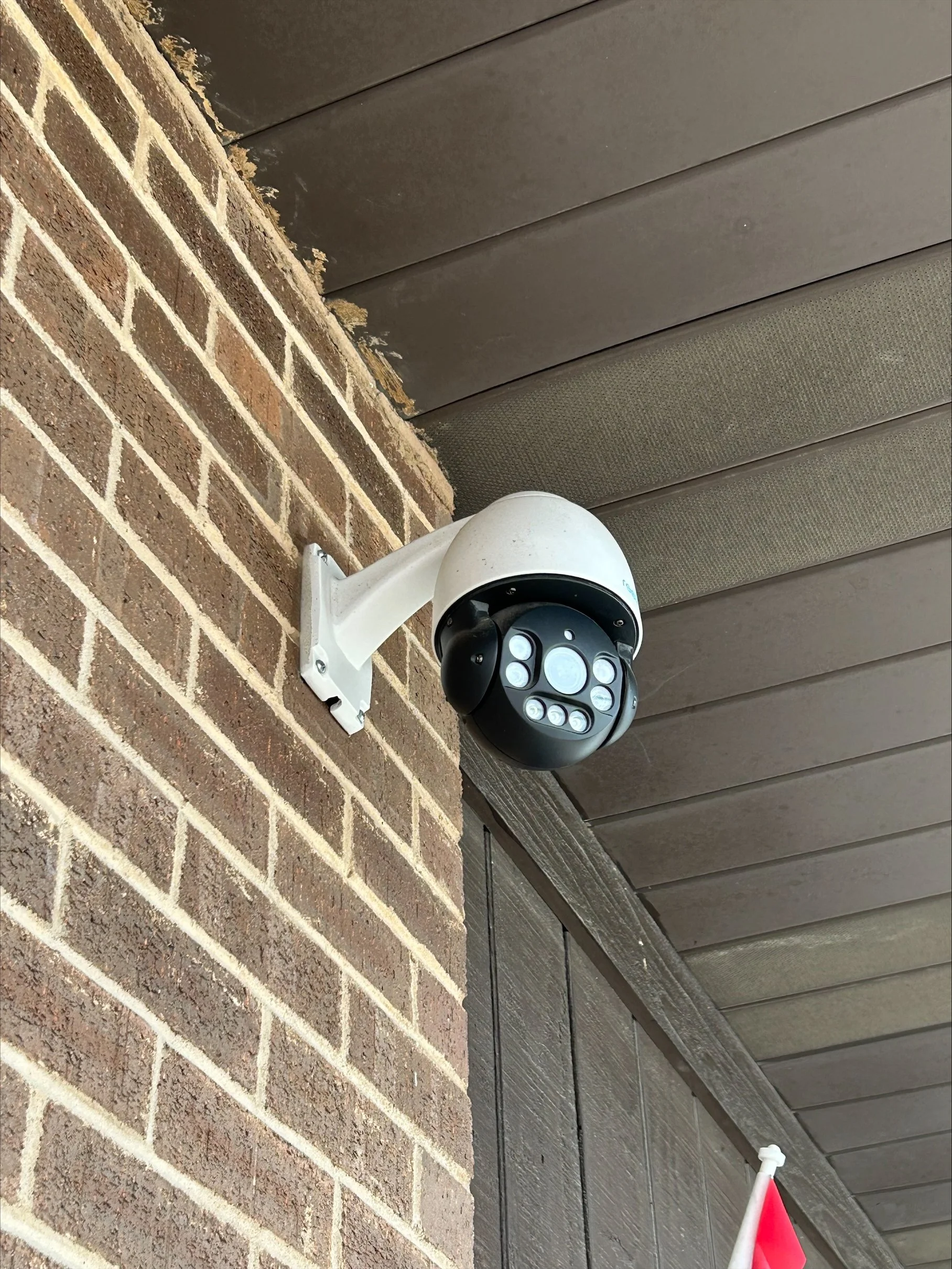 A security camera mounted on a brick wall under a brown wooden ceiling.
