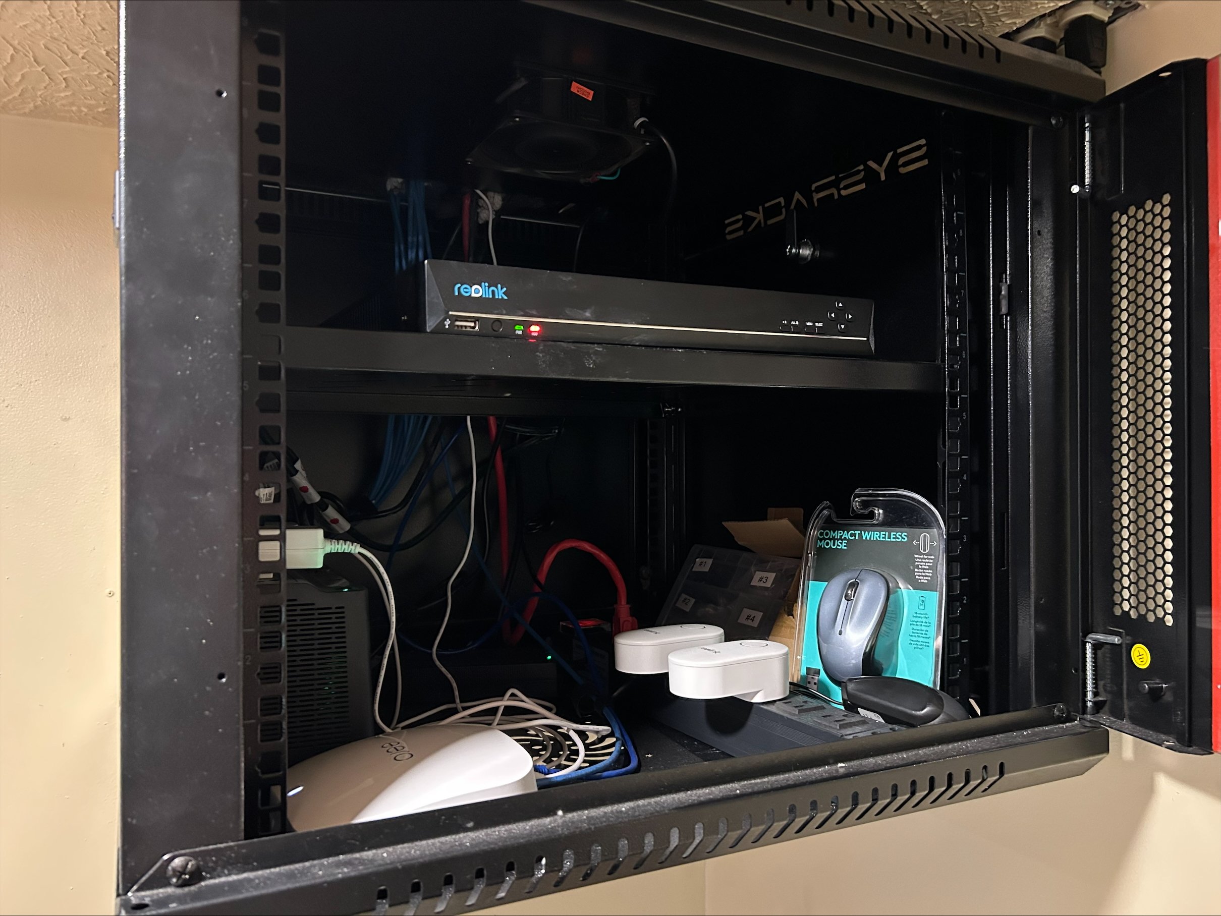 Inside a black metal server rack, various electronic devices, cables, and accessories are arranged, including a Reolink network device, a compact wireless mouse in packaging, and related wiring and power supplies.