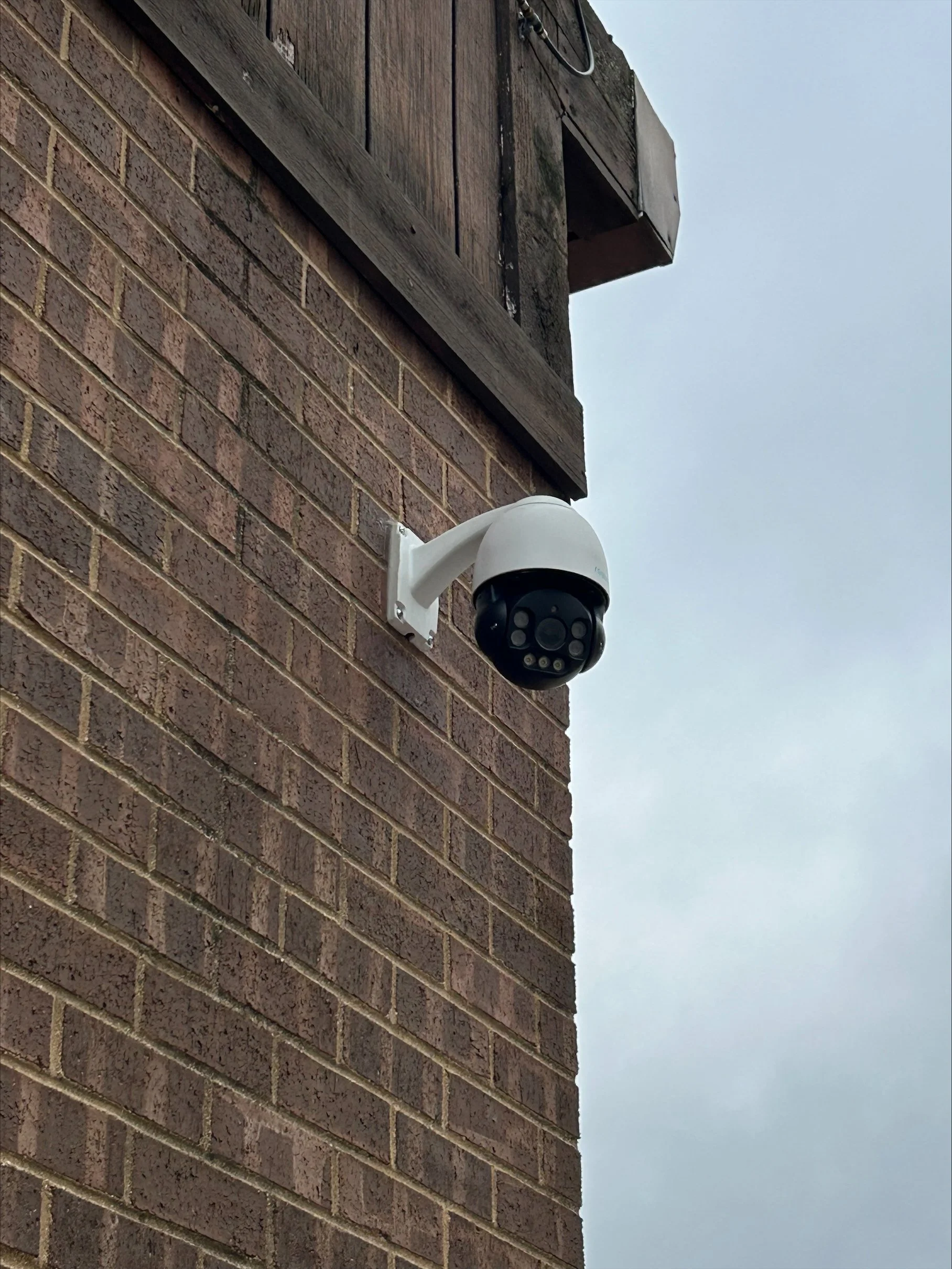 Security camera mounted on the corner of a brick building outdoors under an overcast sky.