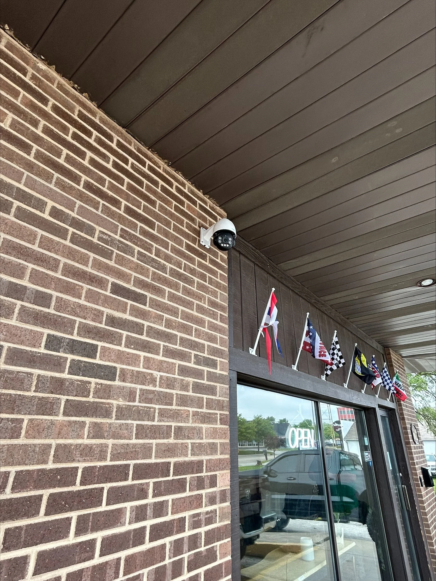 A security camera mounted on the brick wall of a building near the ceiling, with a glass door underneath. Above the door, there are several flags including the American flag and checkered racing flags. An "OPEN" sign is visible through the glass door, along with parked cars and trees outside.
