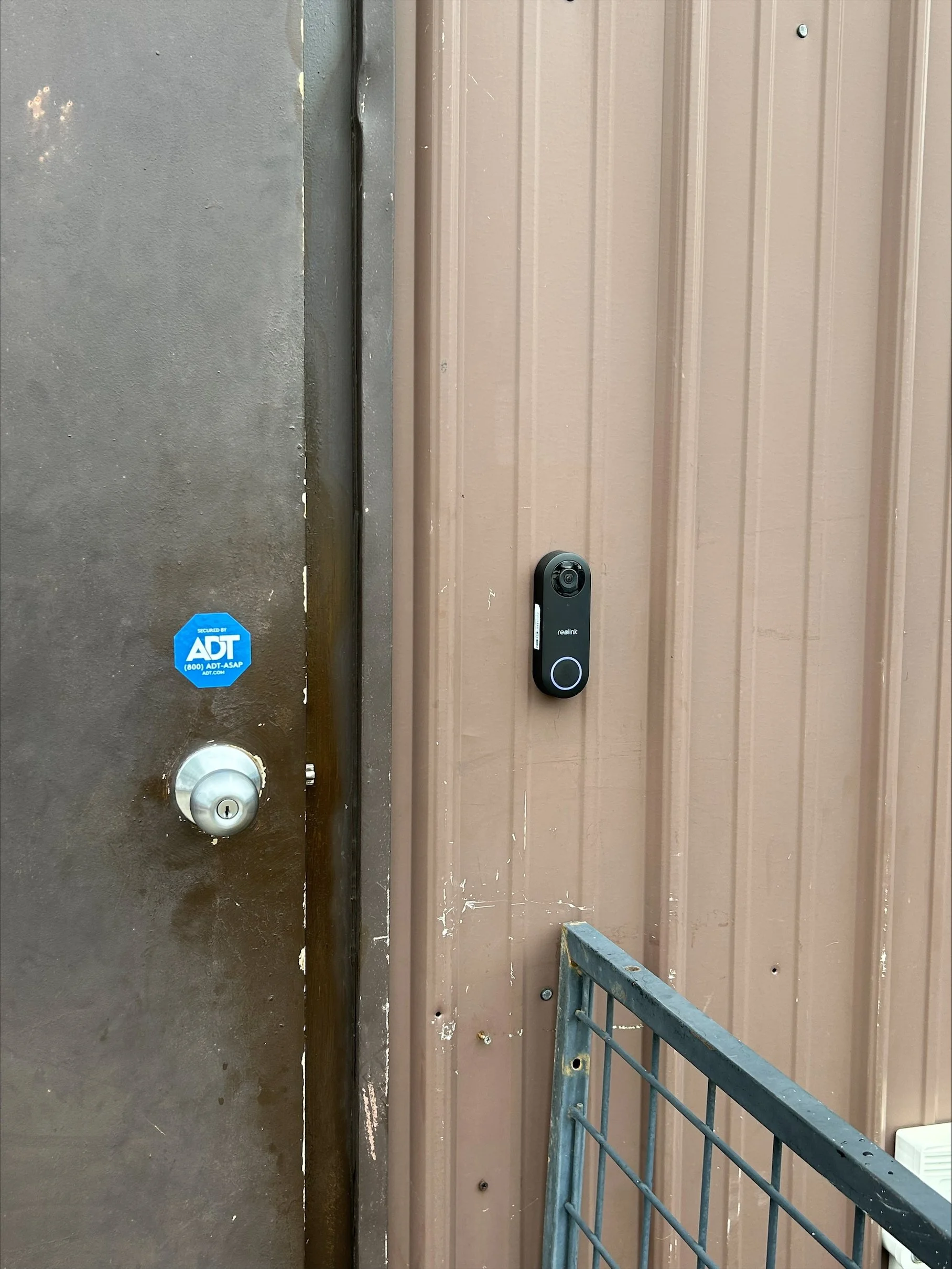 An exterior door with a digital lock and a brown metal wall panel. A small metal gate is visible at the bottom right corner.